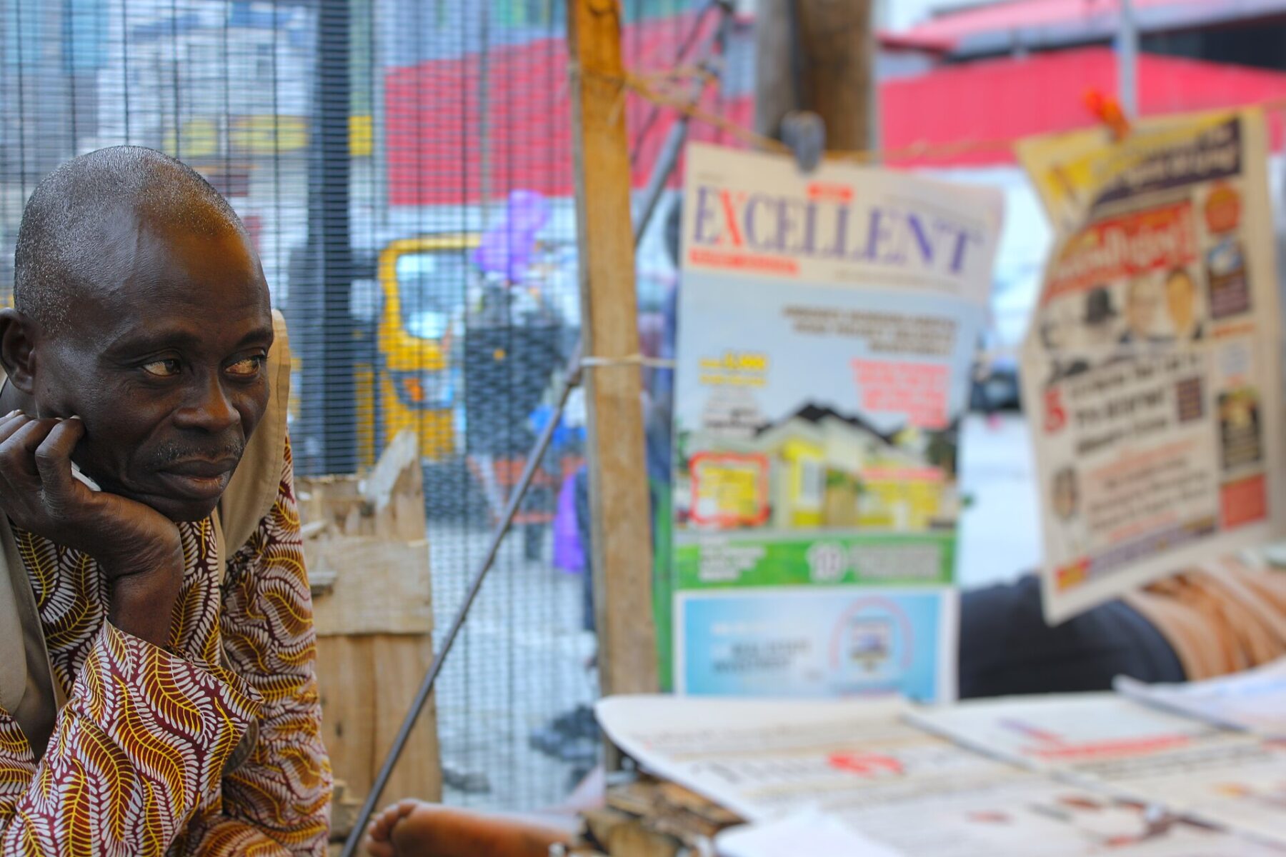 A man in a patterned shirt rests his chin on his hand beside a stall of newspapers and magazines in Nigeria
