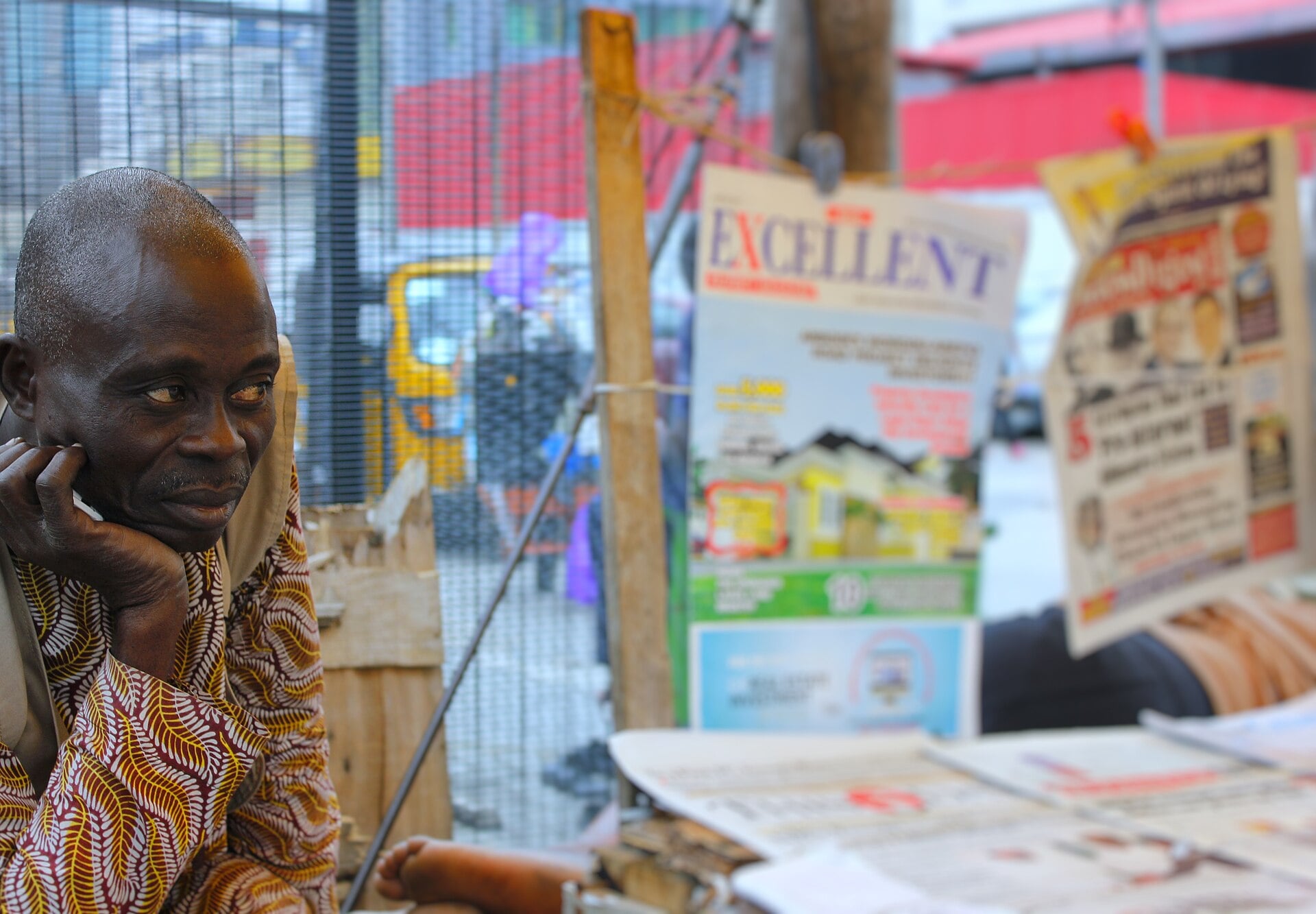 A man in a patterned shirt rests his chin on his hand beside a stall of newspapers and magazines in Nigeria
