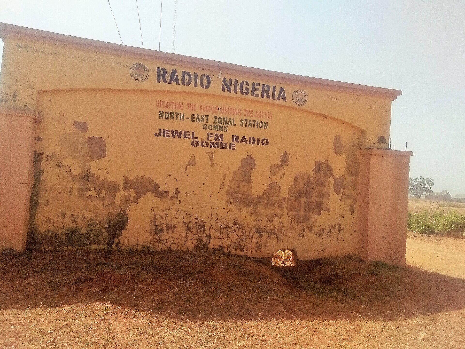 Weathered yellow gate wall reading Radio Nigeria, North-East Zonal Station Gombe, Jewel FM Radio Gombe