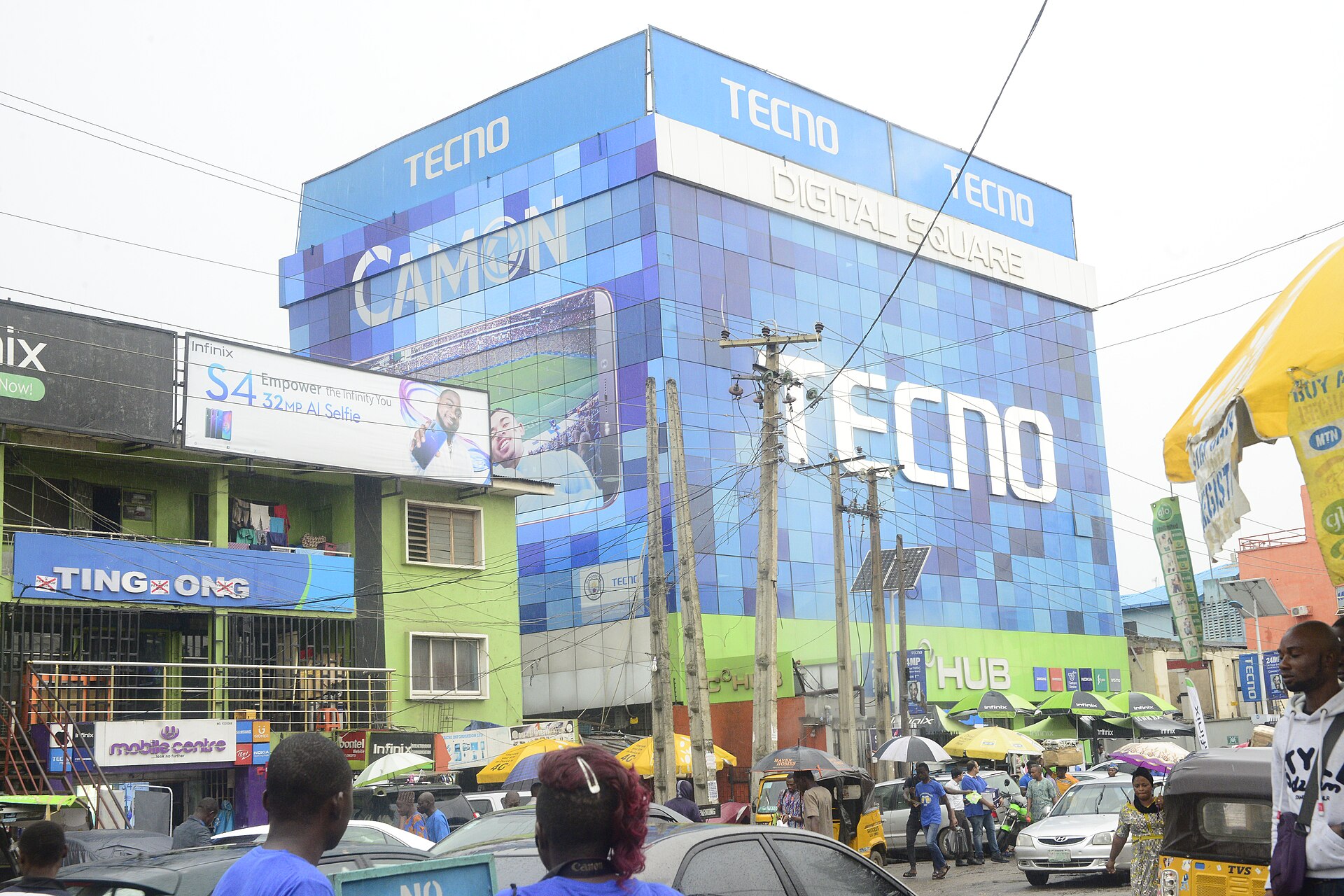 A blue glass building covered in phone brand signs above a busy street with cars, tricycles and pedestrians in Ikeja, Lagos