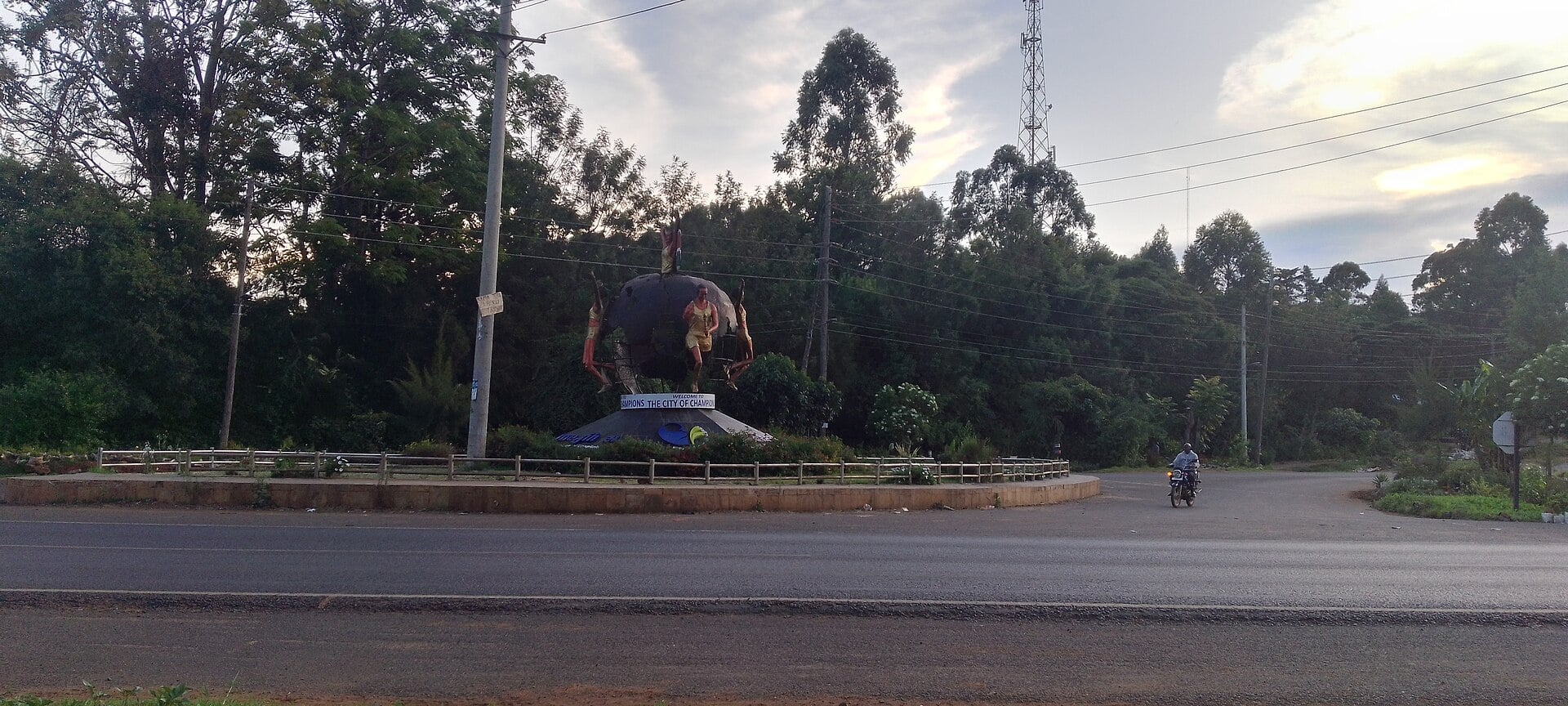 A large globe monument with running figures on a roundabout beside a tree-lined road in Eldoret, with a motorcyclist passing at dusk