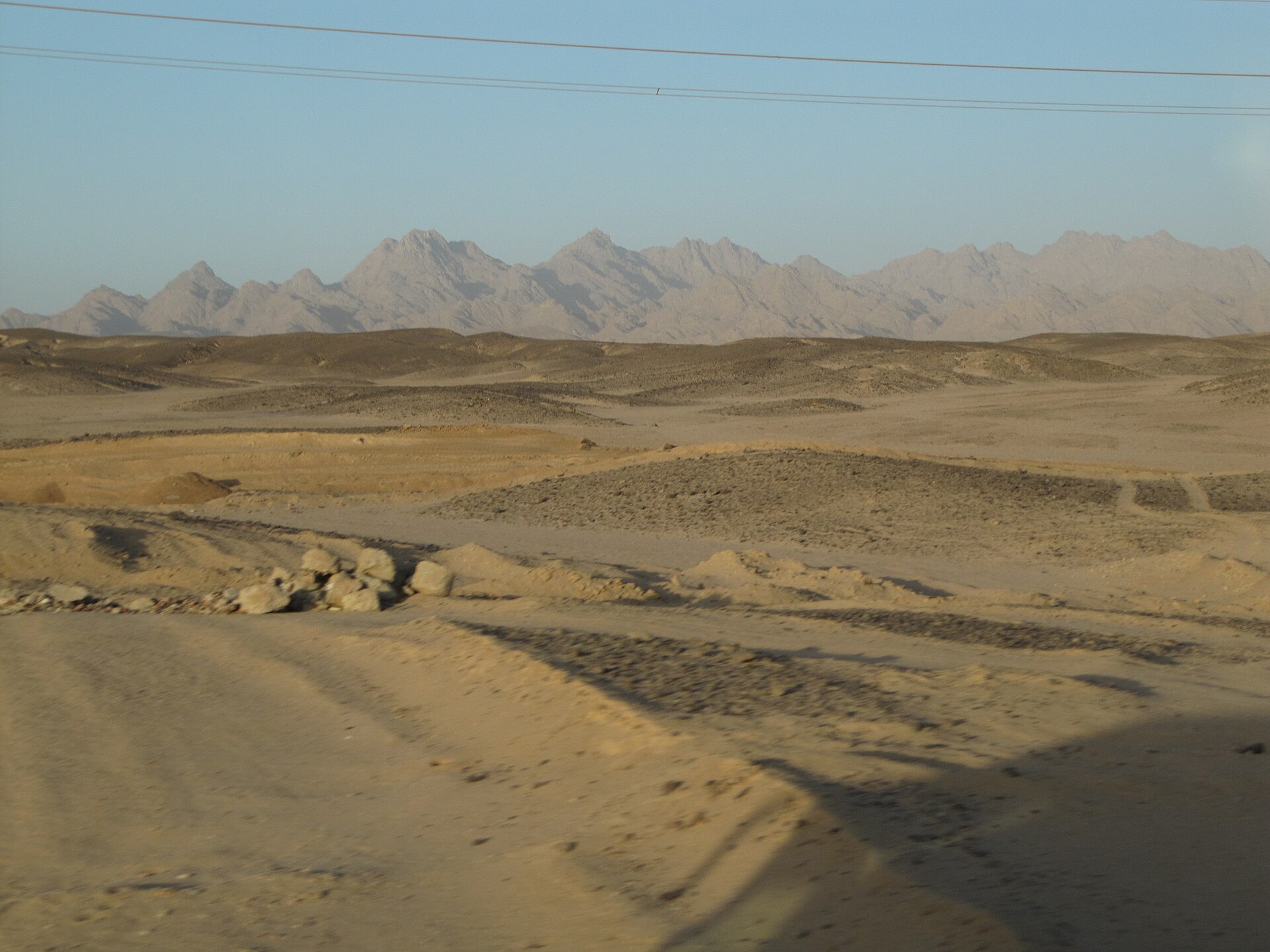 A sandy desert plain with low gravel ridges stretches towards a jagged range of bare grey mountains under a pale sky, with power lines overhead