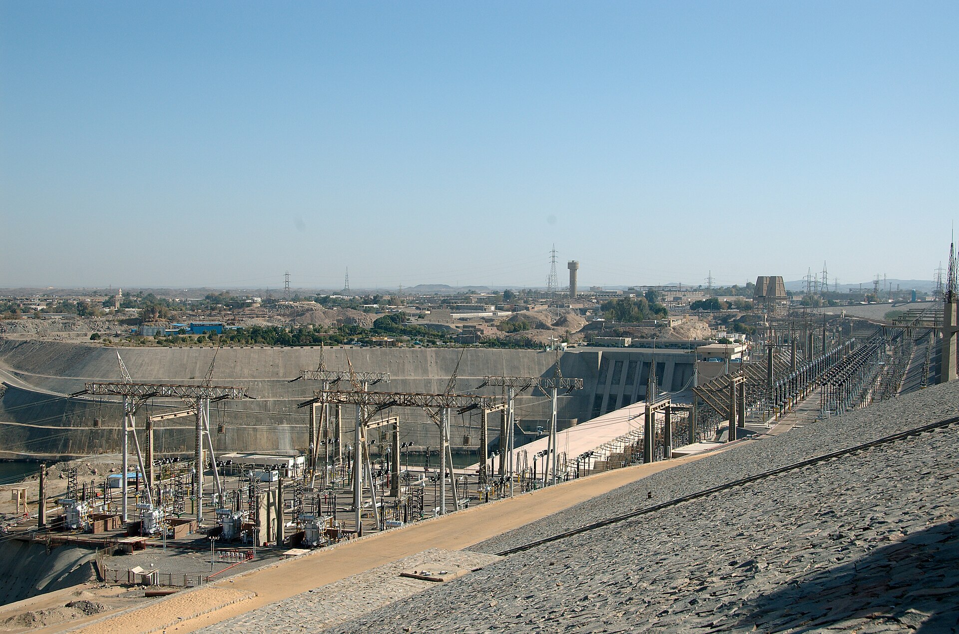 View from the stone-faced crest of the Aswan High Dam down to the hydropower station, its electrical switchyard and pylons, with the town of Aswan behind
