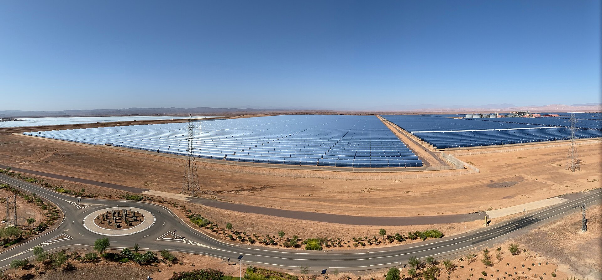 Wide rows of curved solar mirrors stretch across flat desert behind a road roundabout and power pylons under a blue sky