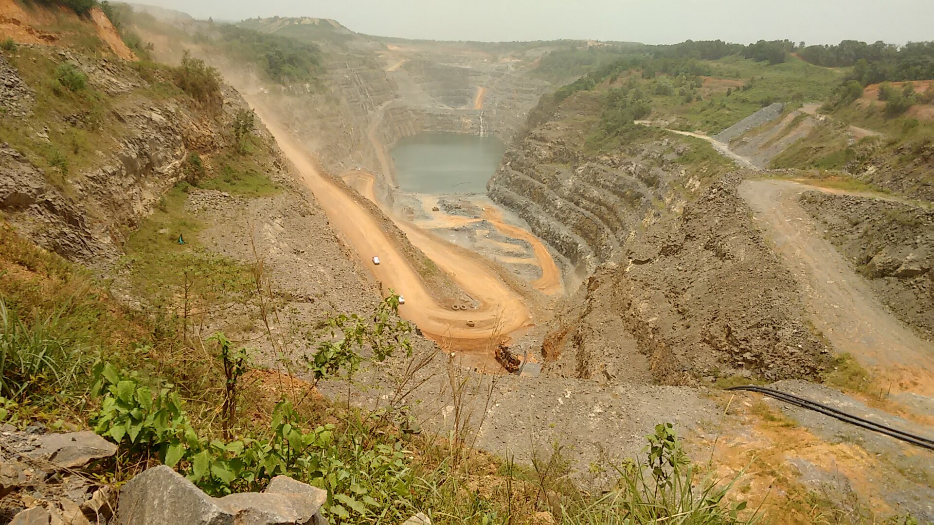 A terraced open-pit gold mine with orange haul roads winding down to a pool of water at the bottom, framed by green scrub and hills