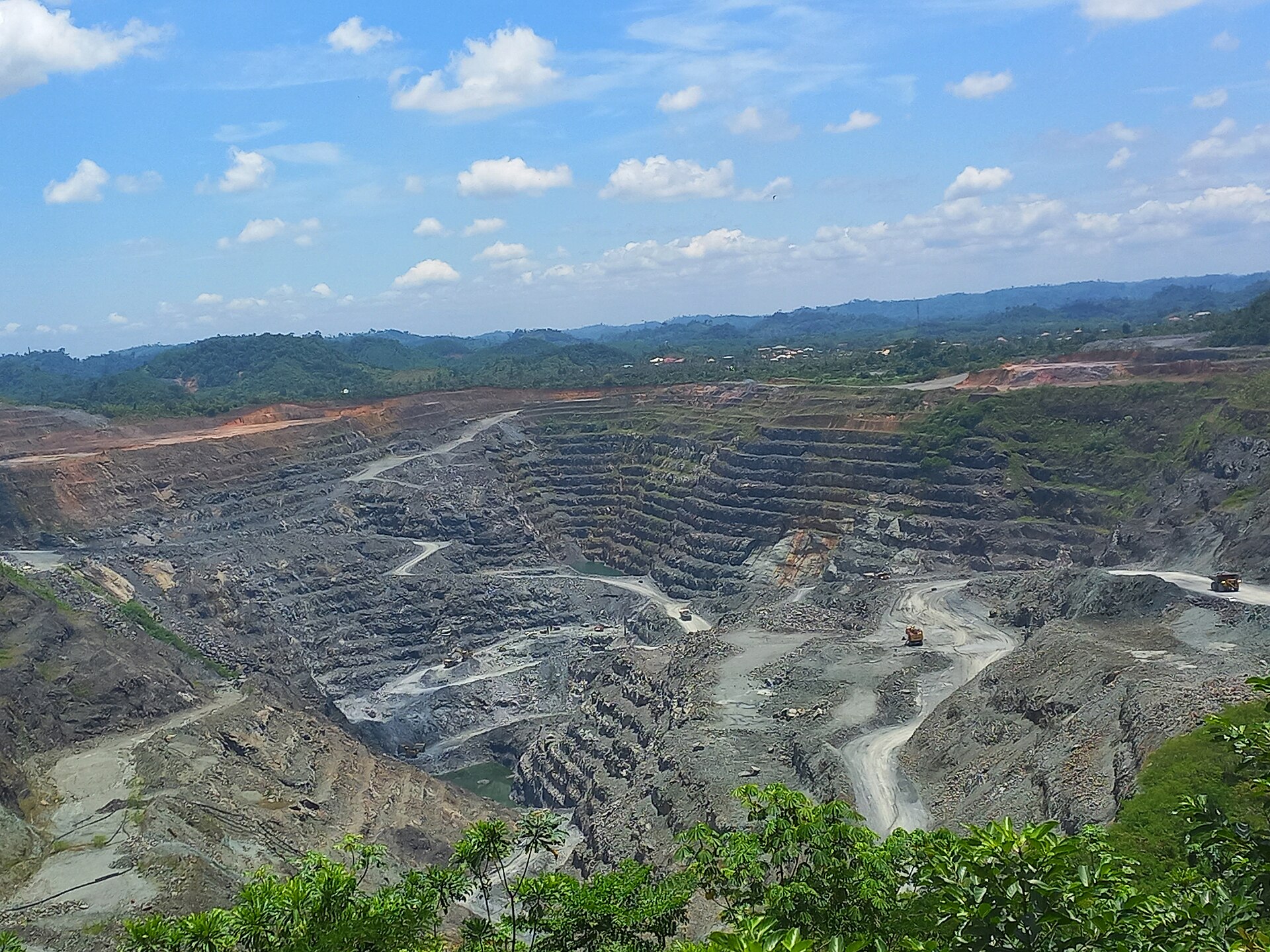 A large terraced open-pit mine of grey rock with haul roads and two trucks, surrounded by green forested hills under a blue sky with white clouds