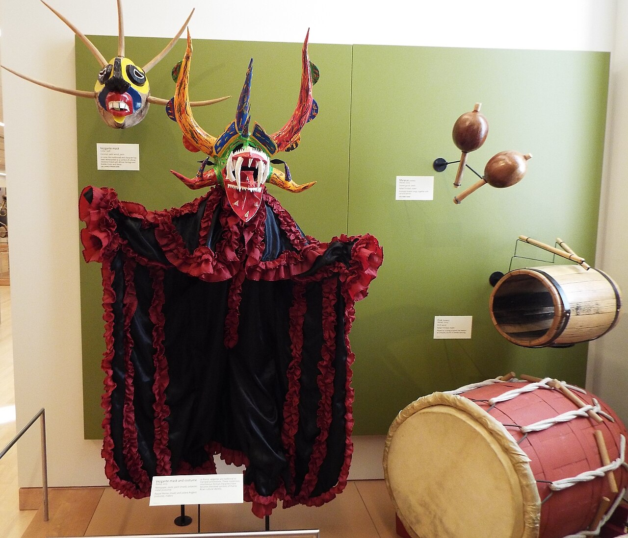A vejigante carnival mask and red-and-black ruffled costume beside maracas and bomba drums in a museum display