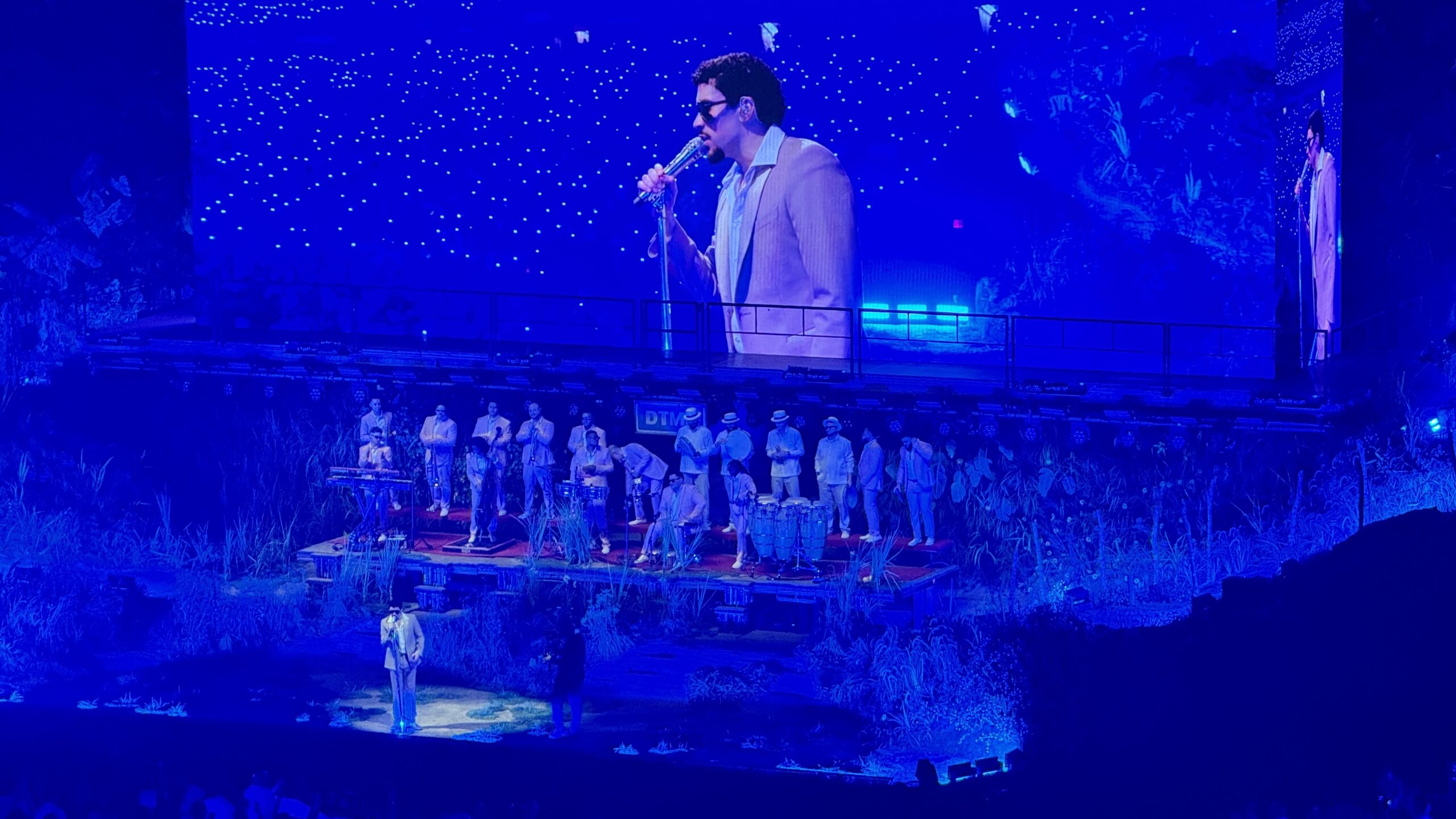 Bad Bunny singing on a blue-lit stage beside a live band with conga drums, with his image on a large screen