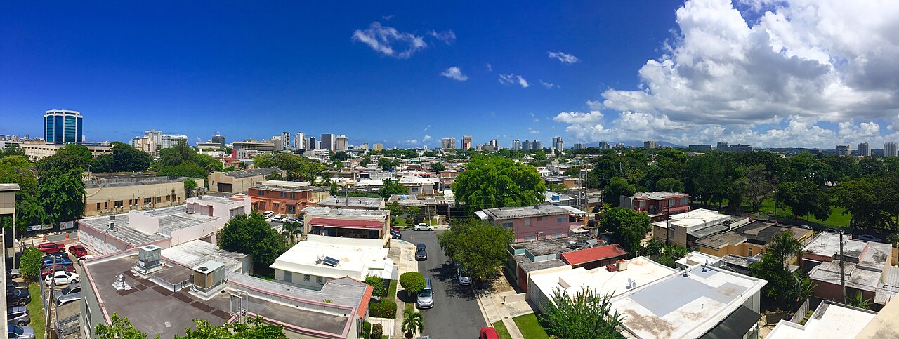 Panoramic view of San Juan's skyline over residential rooftops under a blue sky, with office towers on the horizon