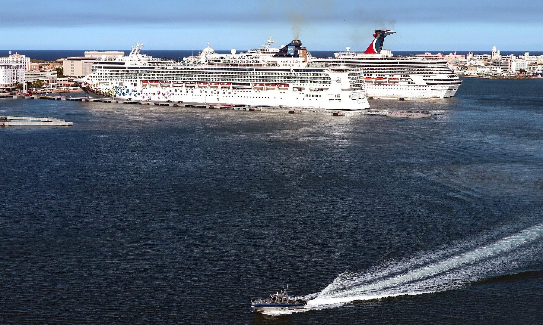 Two cruise ships docked at the San Juan cruise piers beside Old San Juan, with a small boat crossing the harbour in the foreground