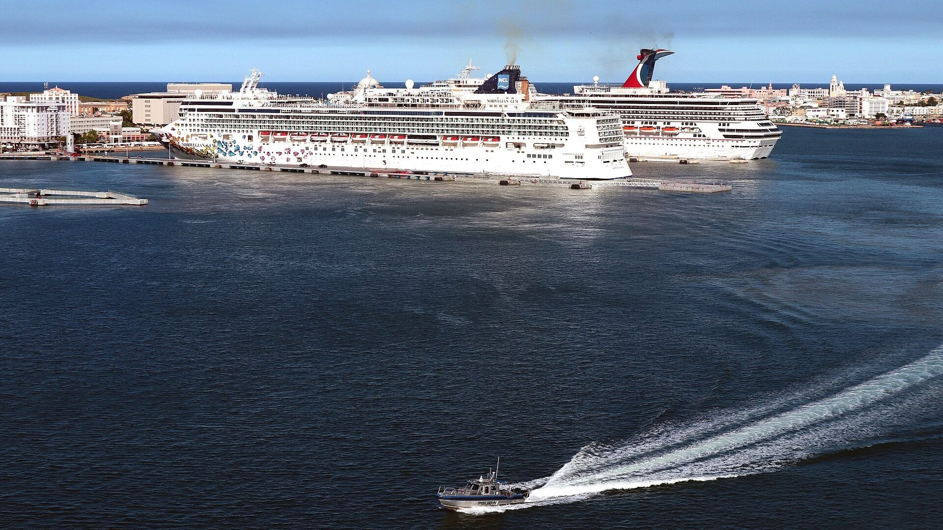 Two cruise ships docked at the San Juan cruise piers beside Old San Juan, with a small boat crossing the harbour in the foreground