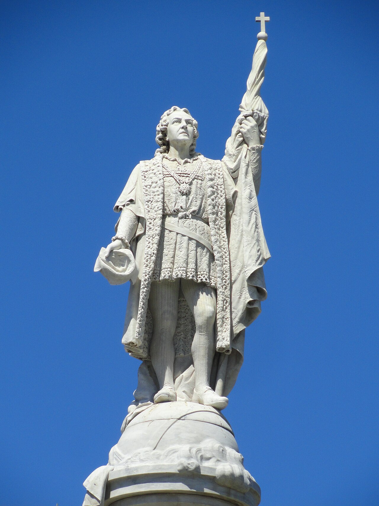 White marble statue of Christopher Columbus holding a flag topped with a cross against a blue sky.