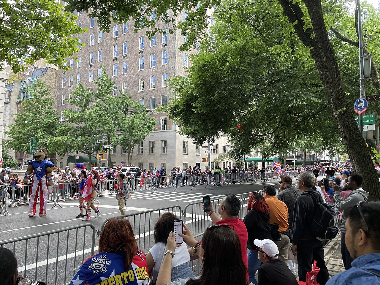 Spectators line a Manhattan avenue to watch the Puerto Rican Day Parade, with Puerto Rican flags and a costumed stilt walker in the street.