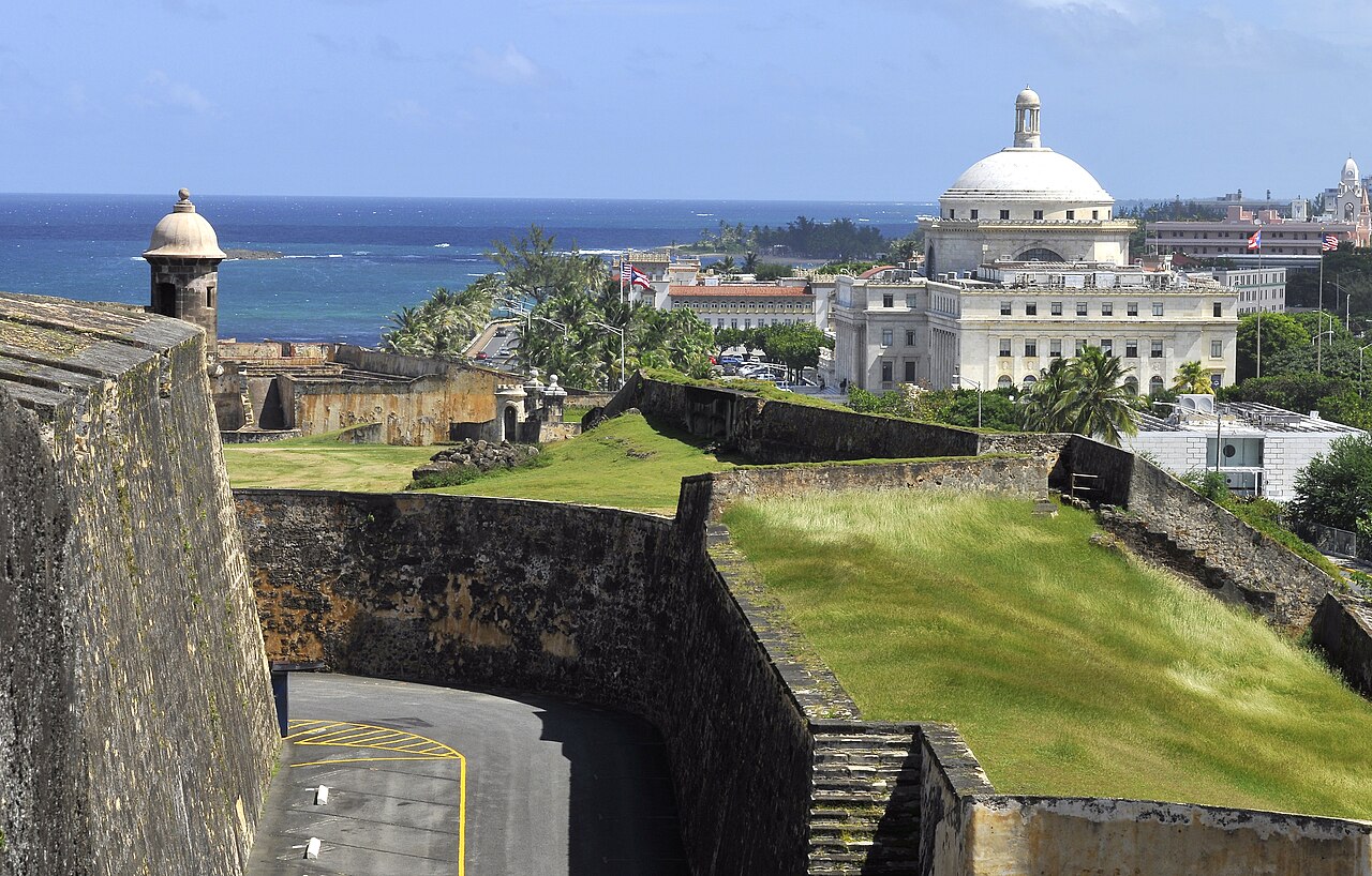 The domed Capitol of Puerto Rico seen from the walls of the San Cristóbal fortress in Old San Juan, with the sea and palm trees beyond.
