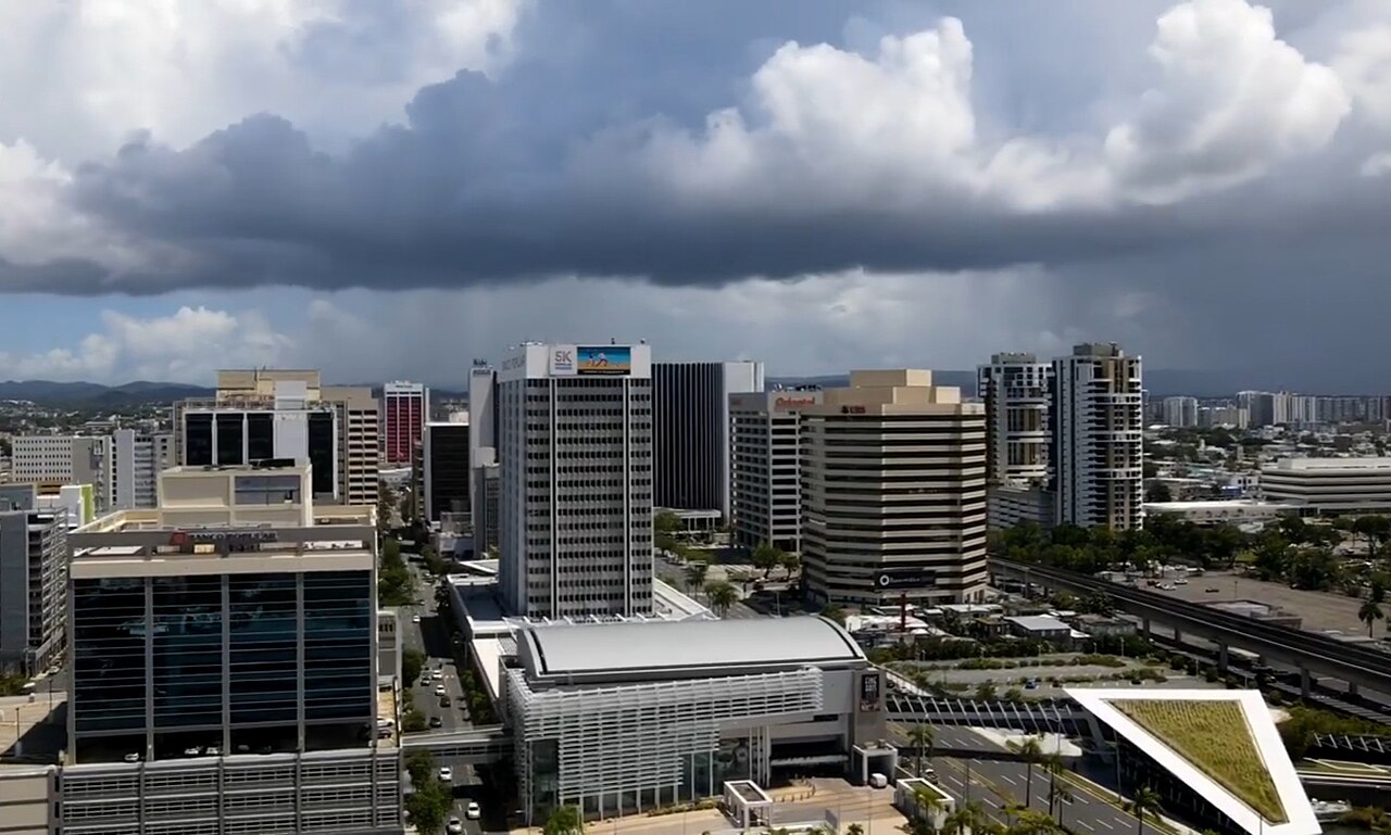 Office towers of the Hato Rey financial district in San Juan under a dark cloudy sky, with a rail viaduct at the right.