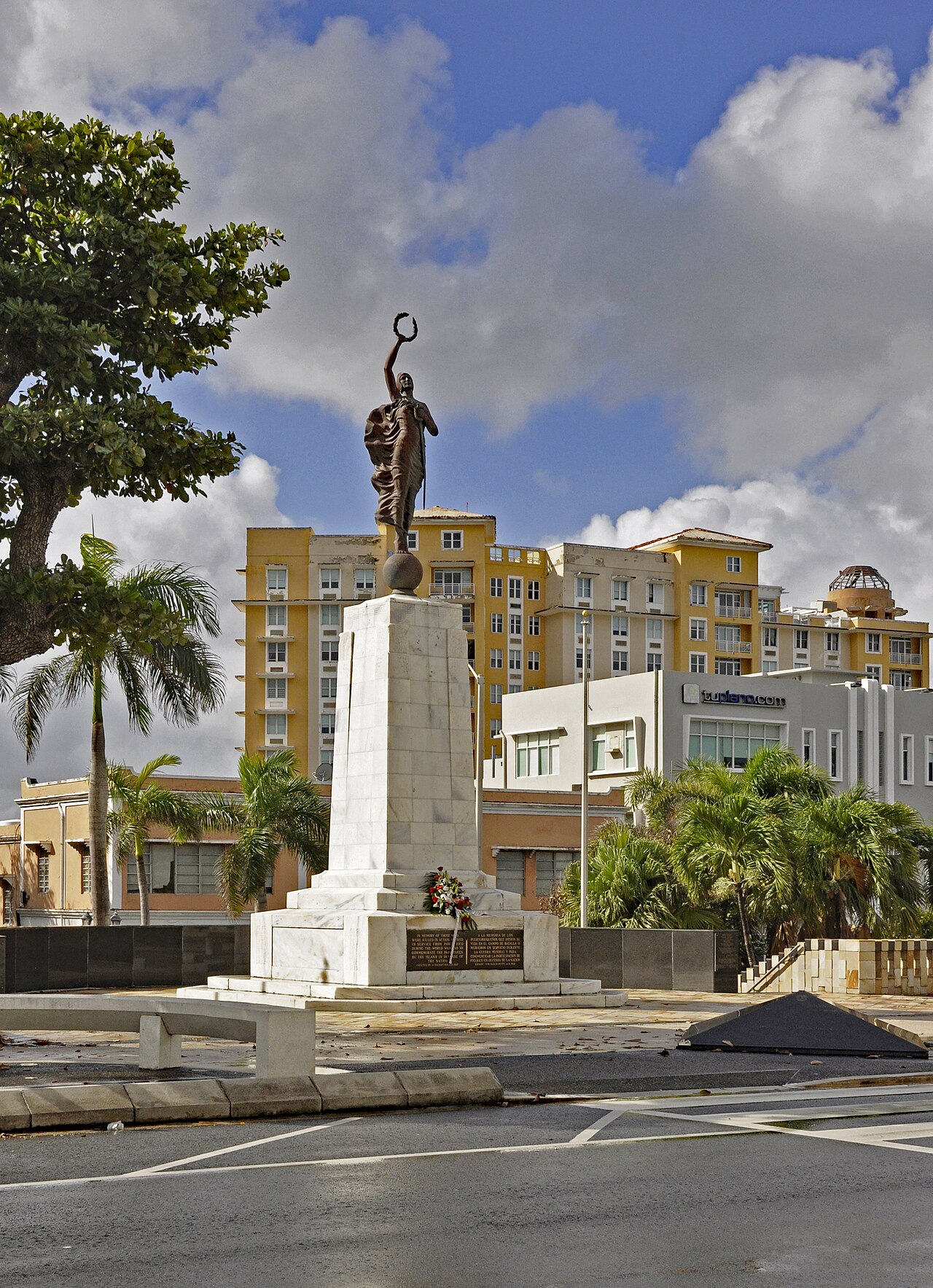 A bronze figure holding a laurel wreath stands on a white marble column in San Juan, with yellow apartment buildings and palm trees behind it.