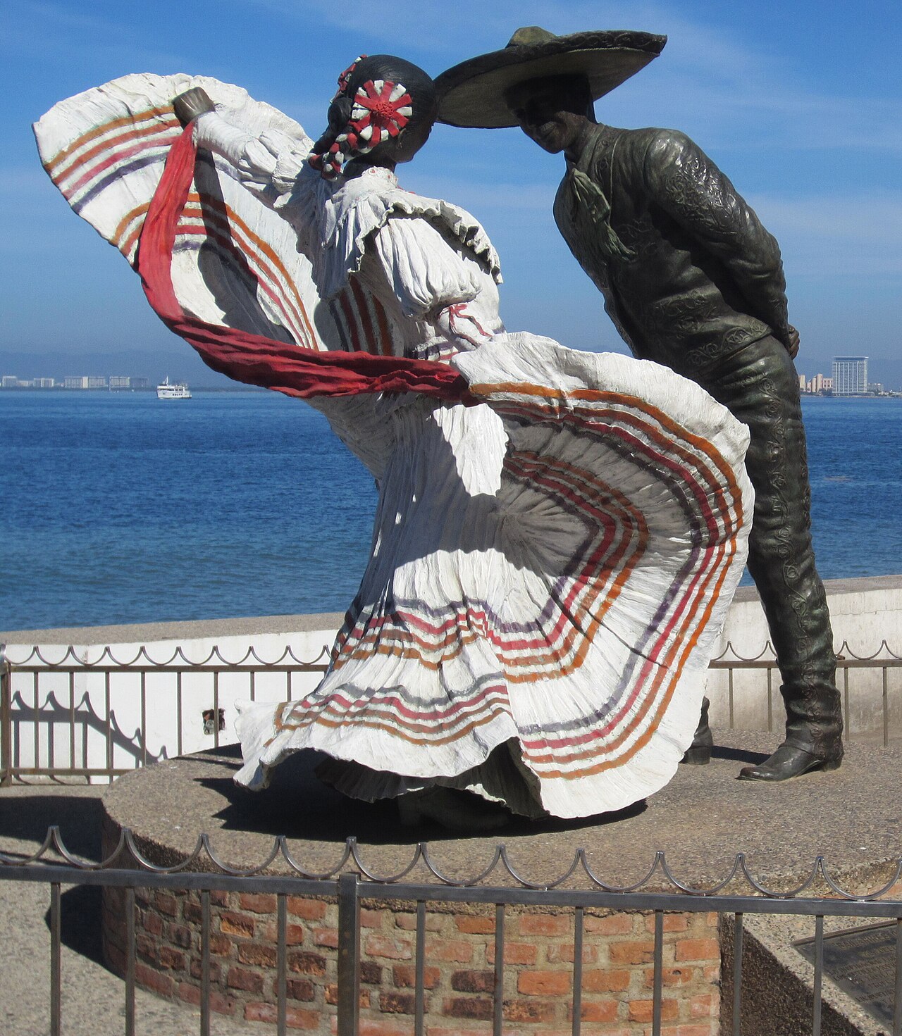 Bronze and painted sculpture of a Mexican folk dancer and a charro in a wide hat on the Malecón promenade, with Banderas Bay behind.