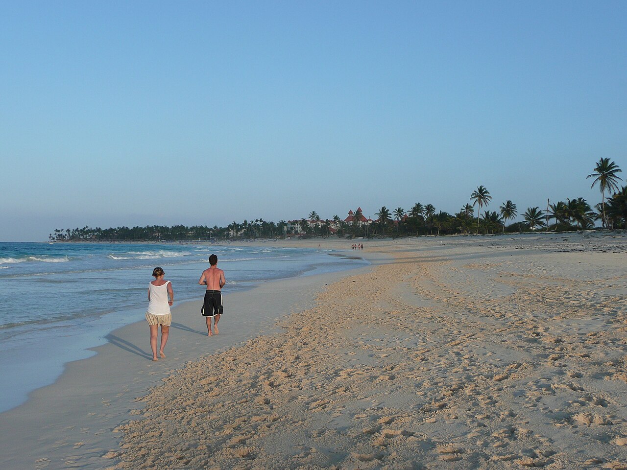 A long sandy beach lined with palm trees and a resort roof in the distance on the coast at Punta Cana, with two people walking by the surf.