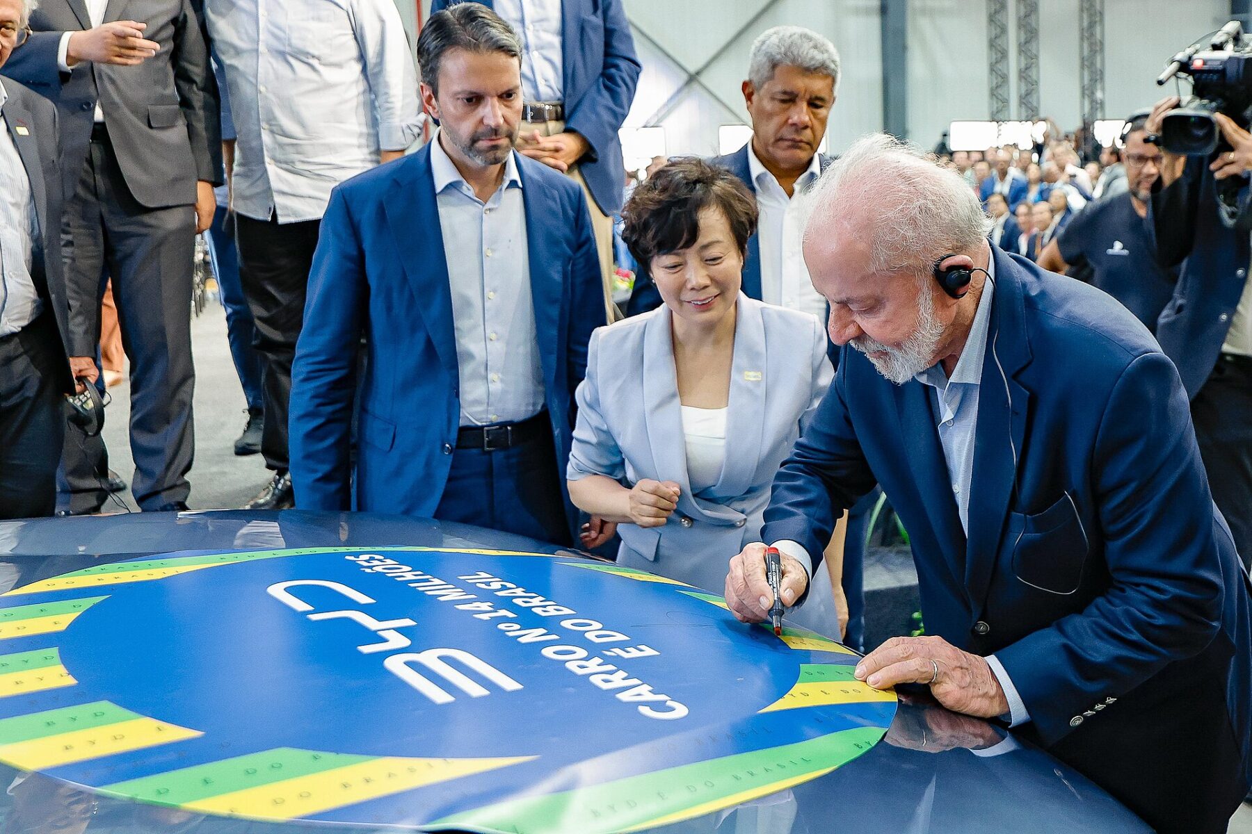 President Lula, wearing an earpiece, signs the bonnet of a car carrying a BYD sticker in Brazil's colours, watched by guests and Brazilian officials