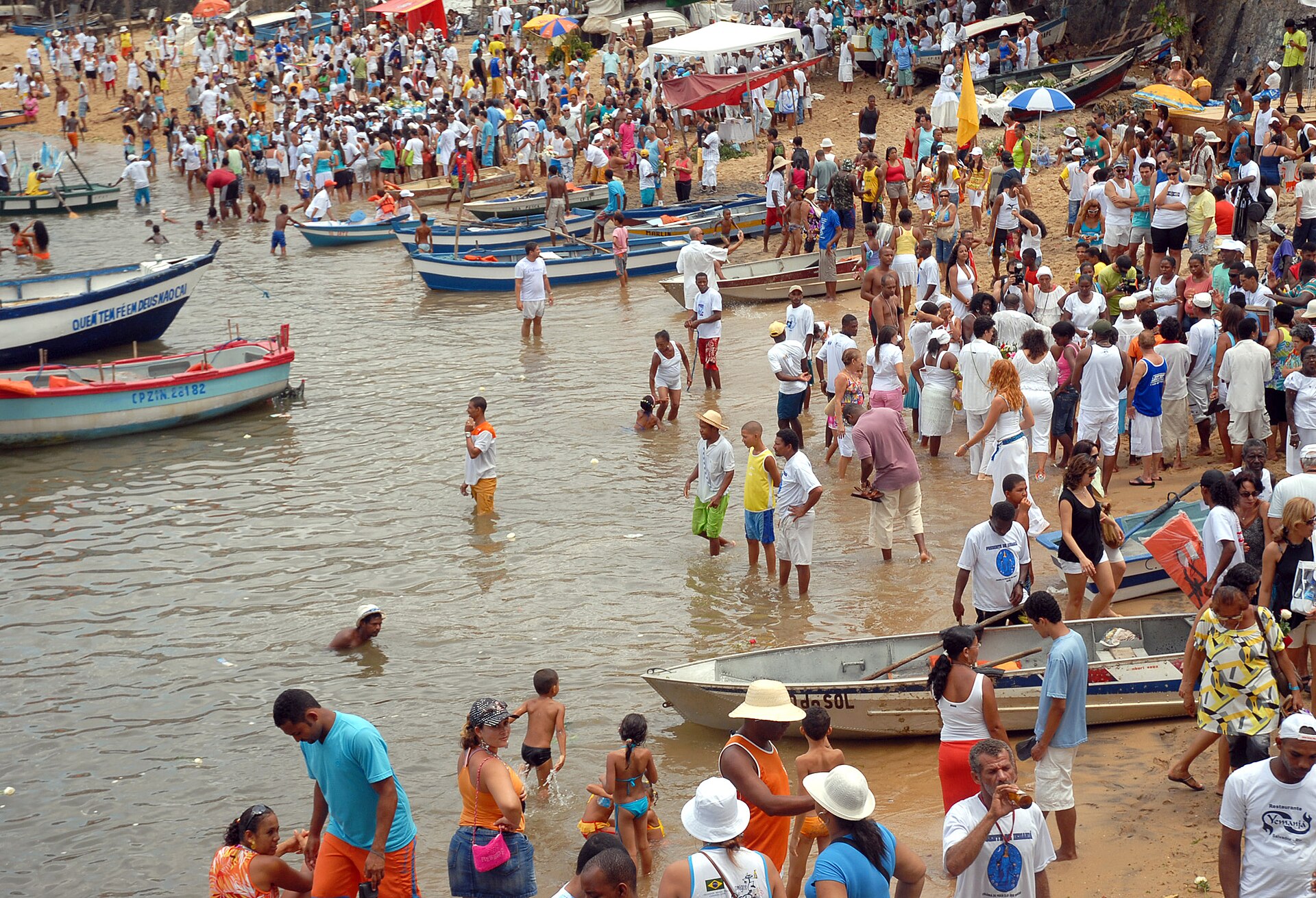 Crowds, many dressed in white, fill a beach and wade into the water among small wooden boats