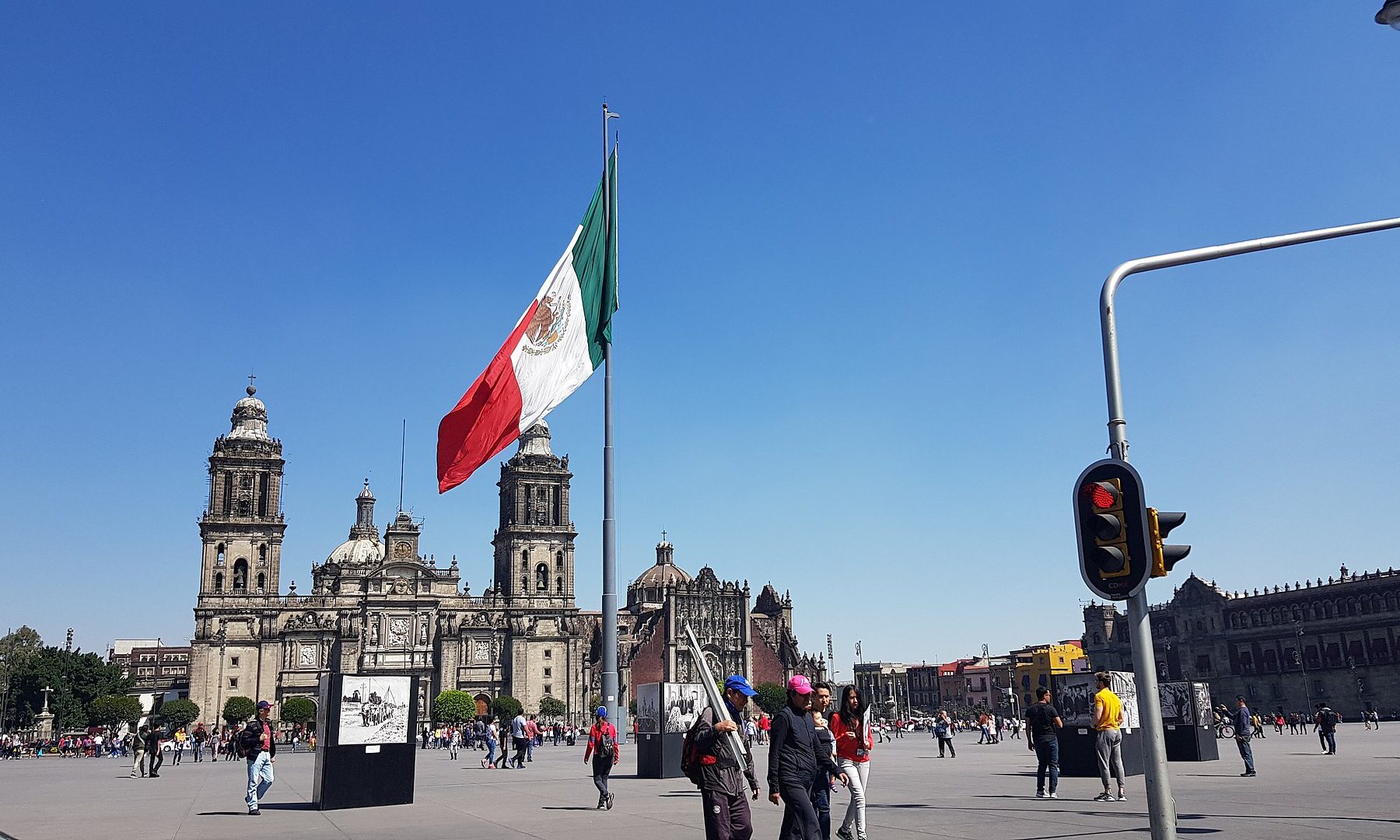 Mexico City's Zócalo square under a blue sky, with a giant Mexican flag, the Metropolitan Cathedral on the left and the National Palace on the right