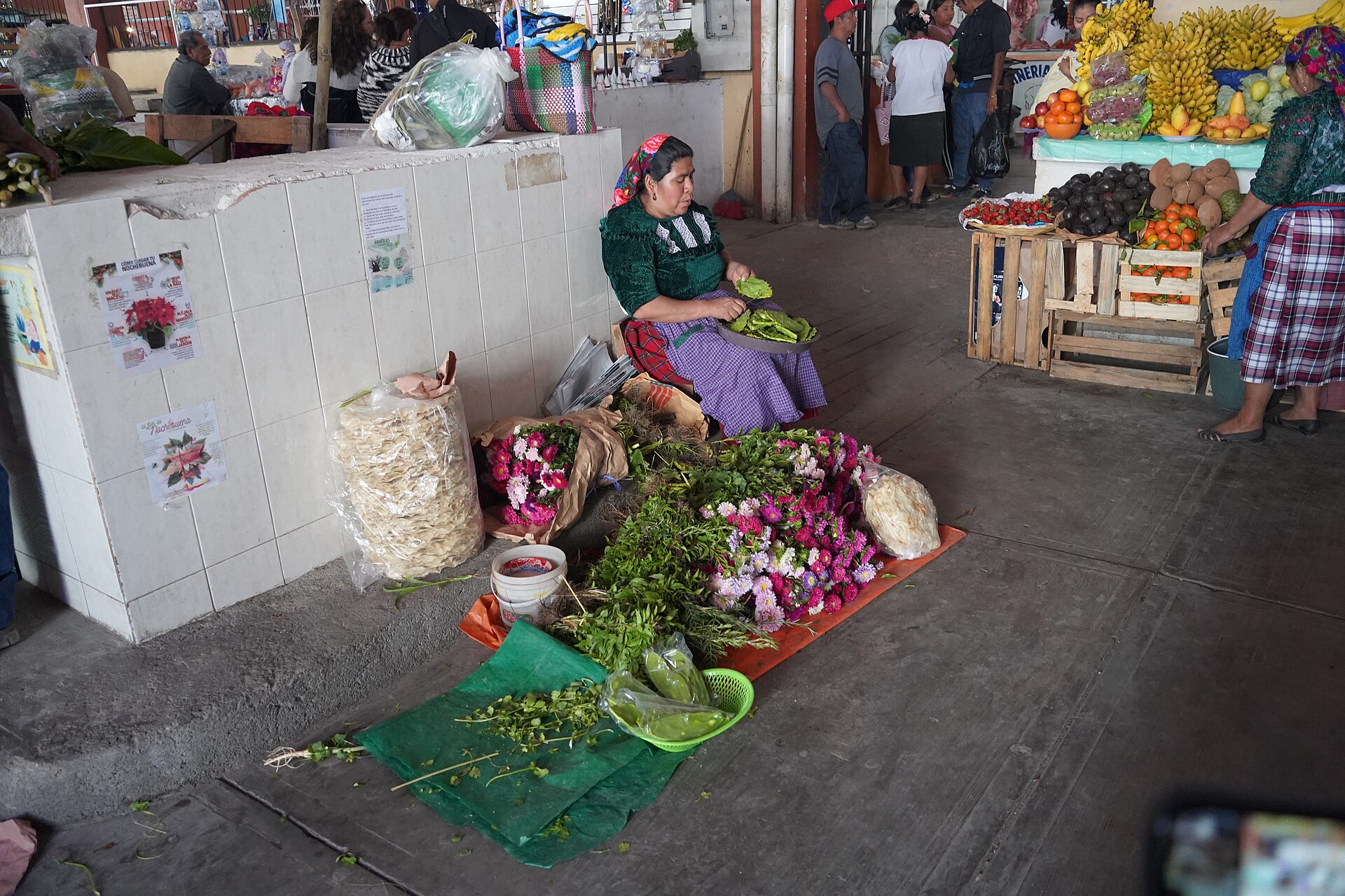 A woman in a green embroidered blouse and headscarf sits on the floor of a covered market beside bundles of flowers and herbs laid out on plastic sheets