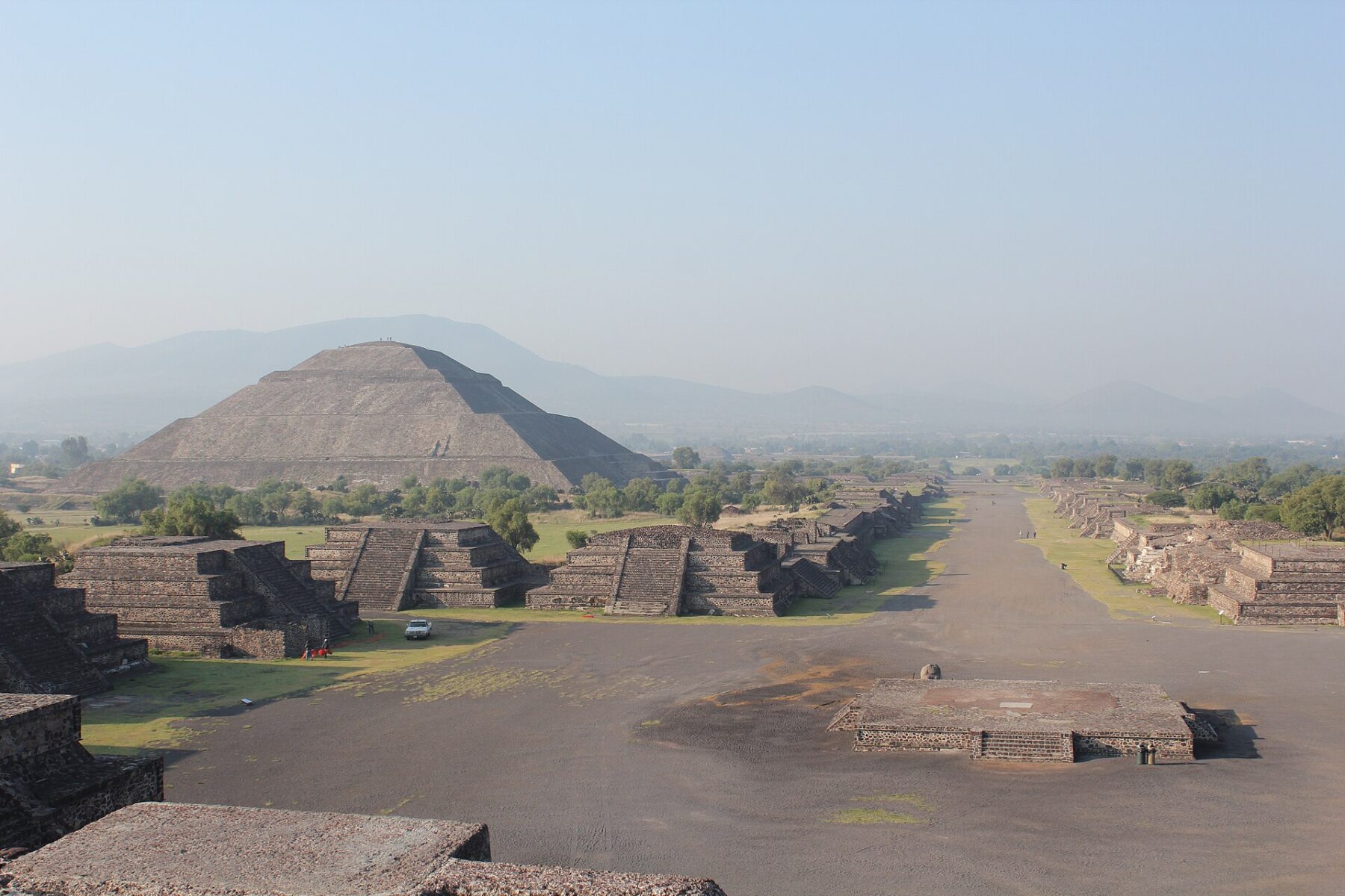 A wide stone avenue lined with low stepped platforms leads toward the large Pyramid of the Sun at Teotihuacan, with hazy mountains behind