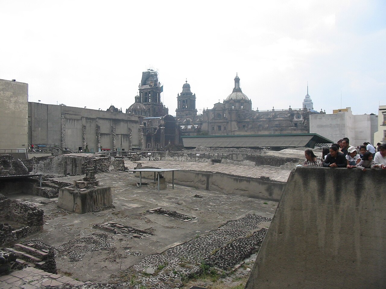 Excavated stone foundations of the Templo Mayor precinct in the foreground, with the domes and bell towers of Mexico City's Metropolitan Cathedral behind and visitors leaning on a wall at right