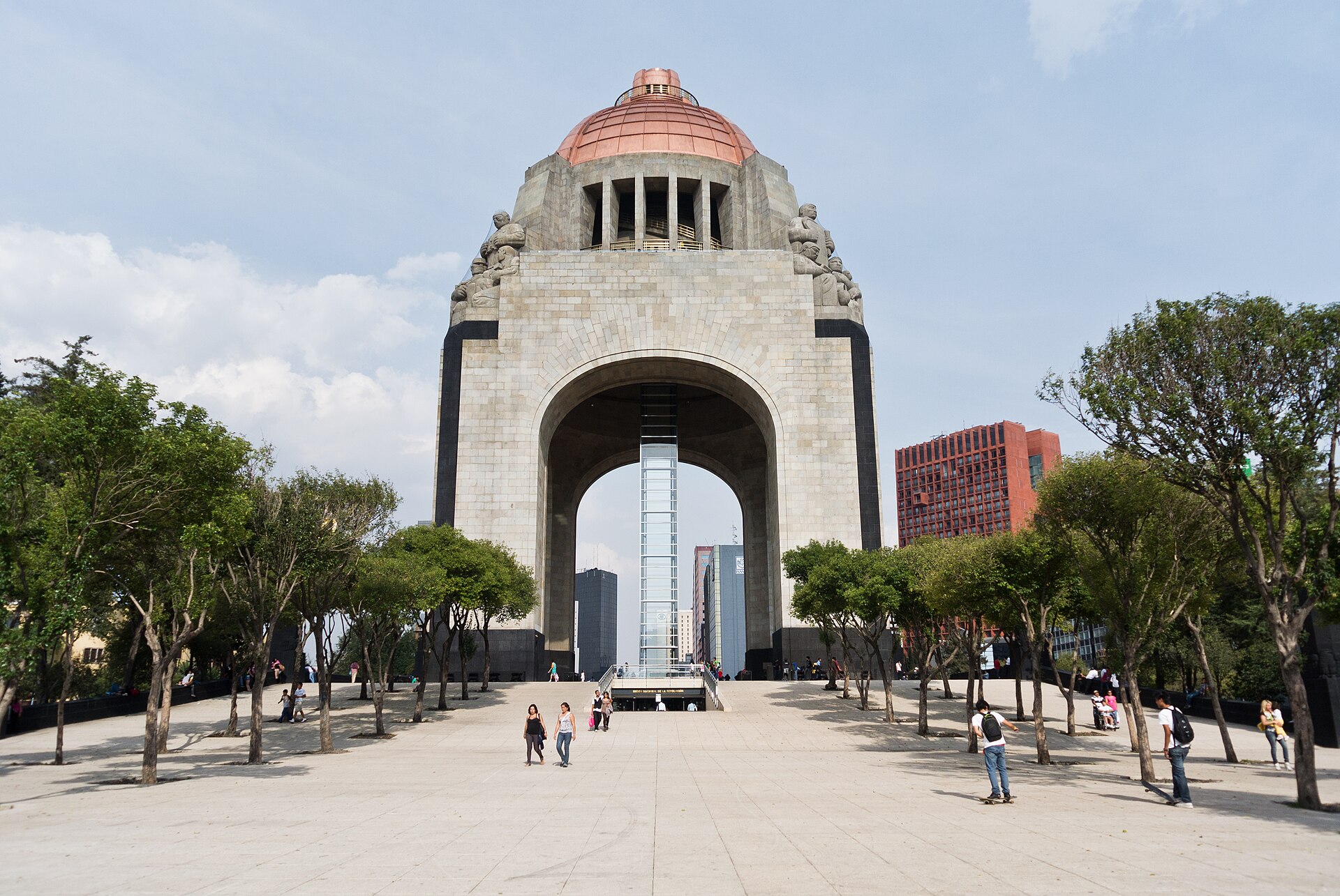 A massive stone arch topped by a copper dome stands at the end of a wide tree-lined plaza, with a few pedestrians walking across