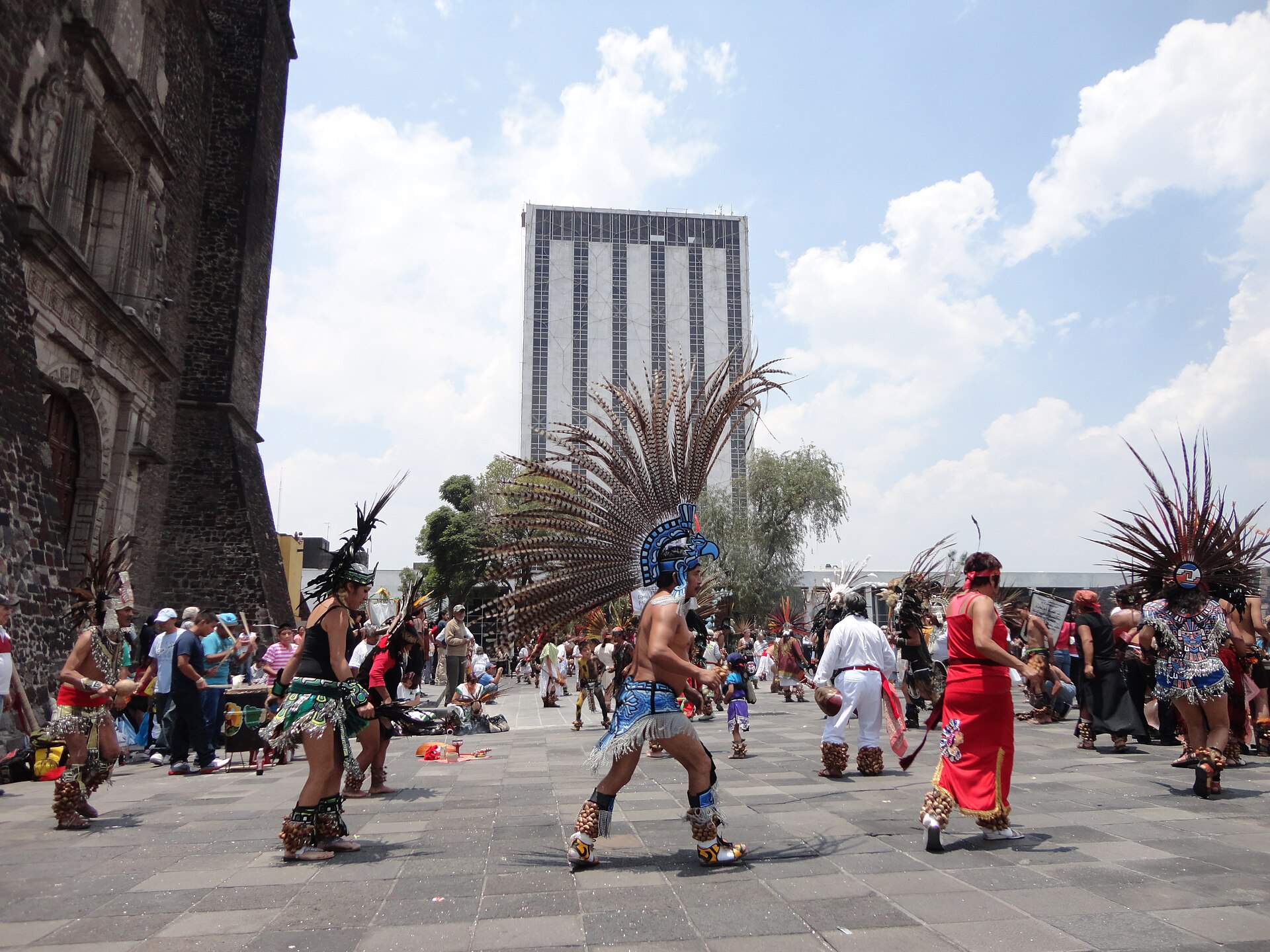 Dancers in tall feathered headdresses and costumes perform on a paved plaza, with a dark stone church at left, a modern high-rise behind and onlookers around them
