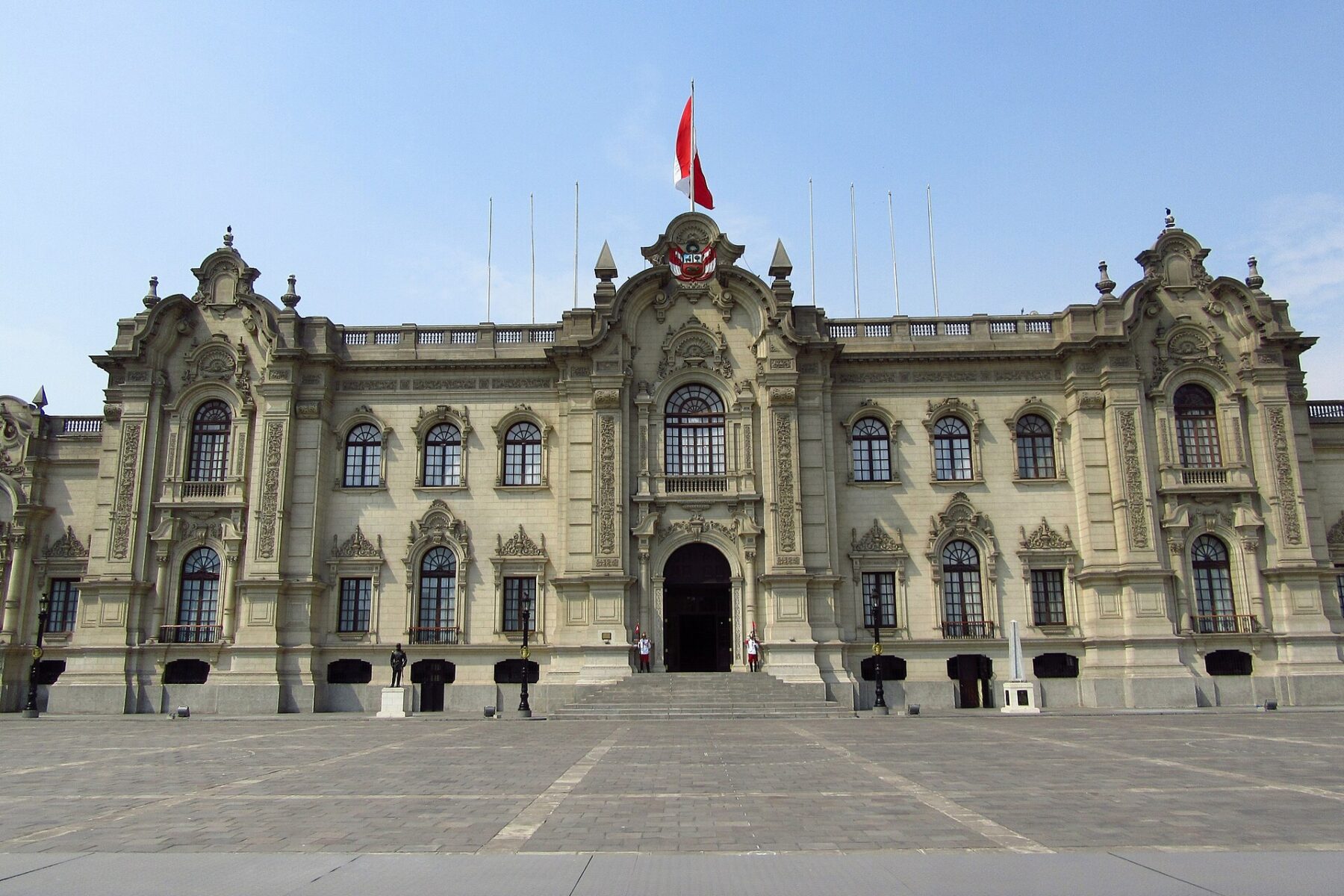 The cream-coloured stone facade of Peru's Government Palace in Lima, with a Peruvian flag on the roof and two guards at the entrance