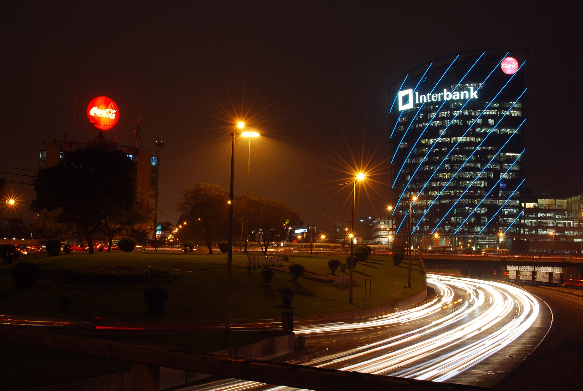 A glass office tower lit with blue diagonal lines and an Interbank sign at night, beside a road with light trails from passing traffic