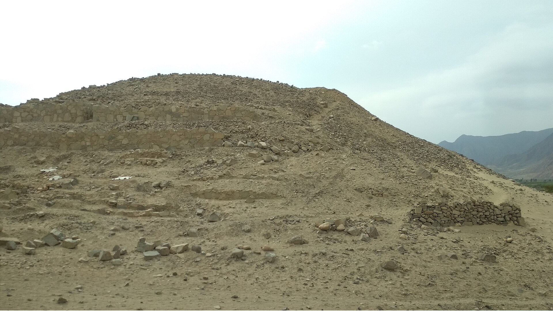 A stepped stone platform mound at the Caral archaeological site in a dry desert valley with mountains in the background