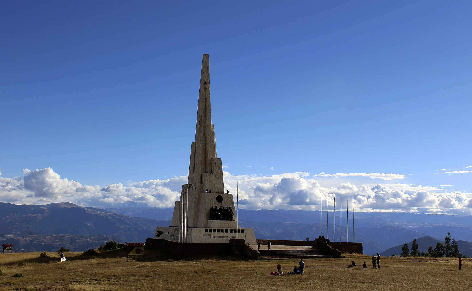 A tall white stone obelisk on a grassy high plain with visitors in front and Andean mountains and clouds behind