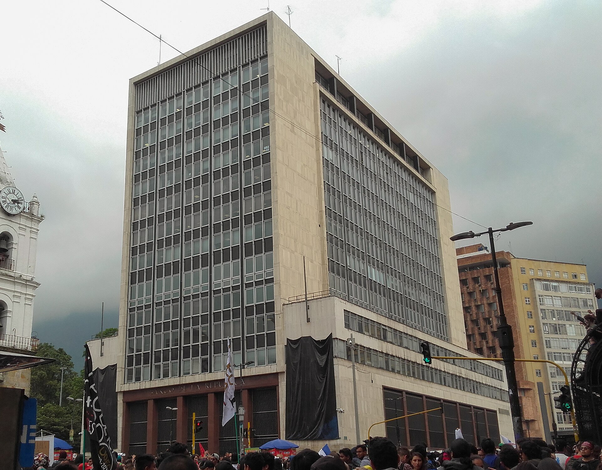 The tall stone-and-glass tower of the Banco de la República in central Bogotá under a grey sky, with a crowd in the street below