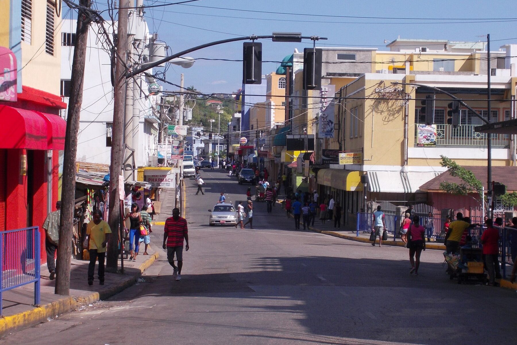 A sunny street in Montego Bay with pedestrians, shops with red and yellow awnings, traffic lights and overhead wires