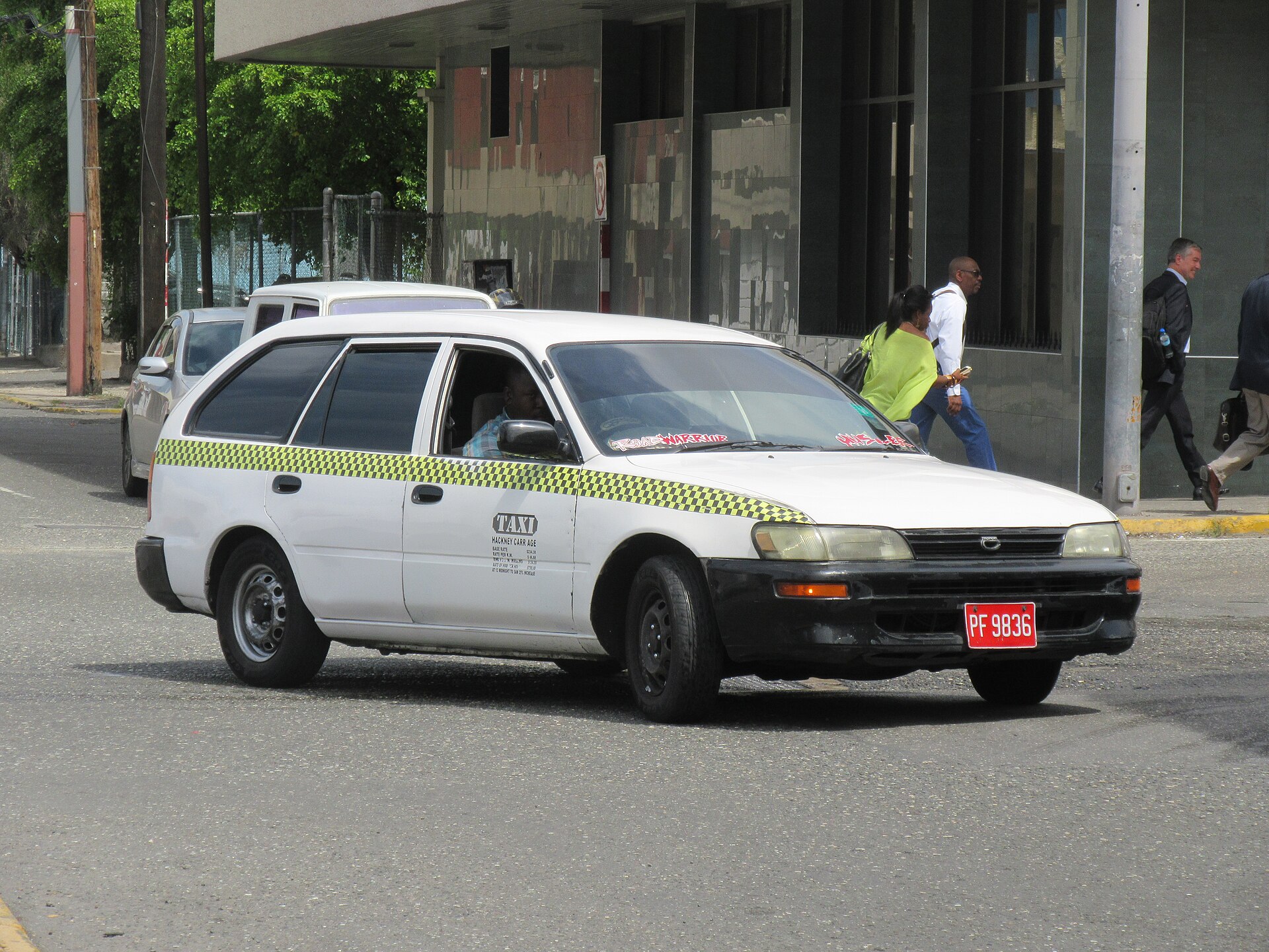 A white Toyota Corolla station wagon taxi with a lime-green and black checked stripe, a TAXI and HACKNEY CARRIAGE sign on the door and a red licence plate