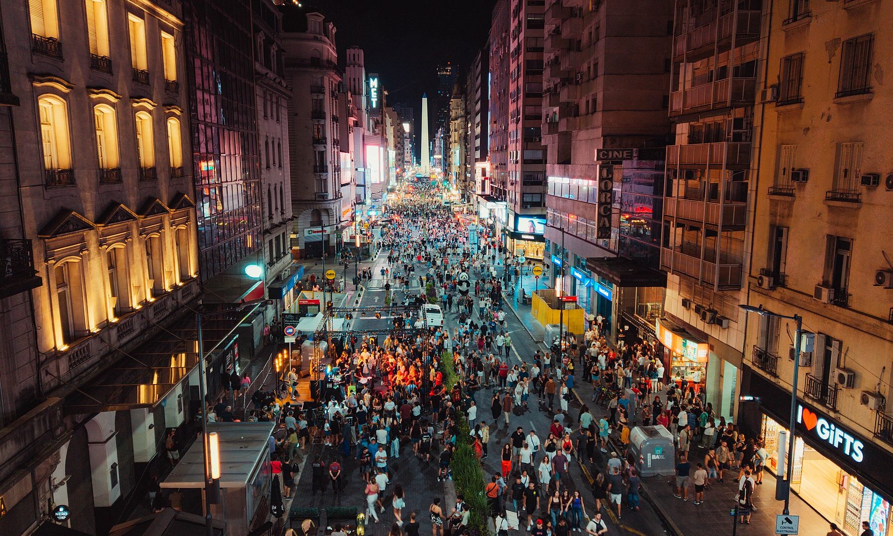 Aerial view of a dense crowd filling a brightly lit avenue at night, with the Obelisk of Buenos Aires in the distance