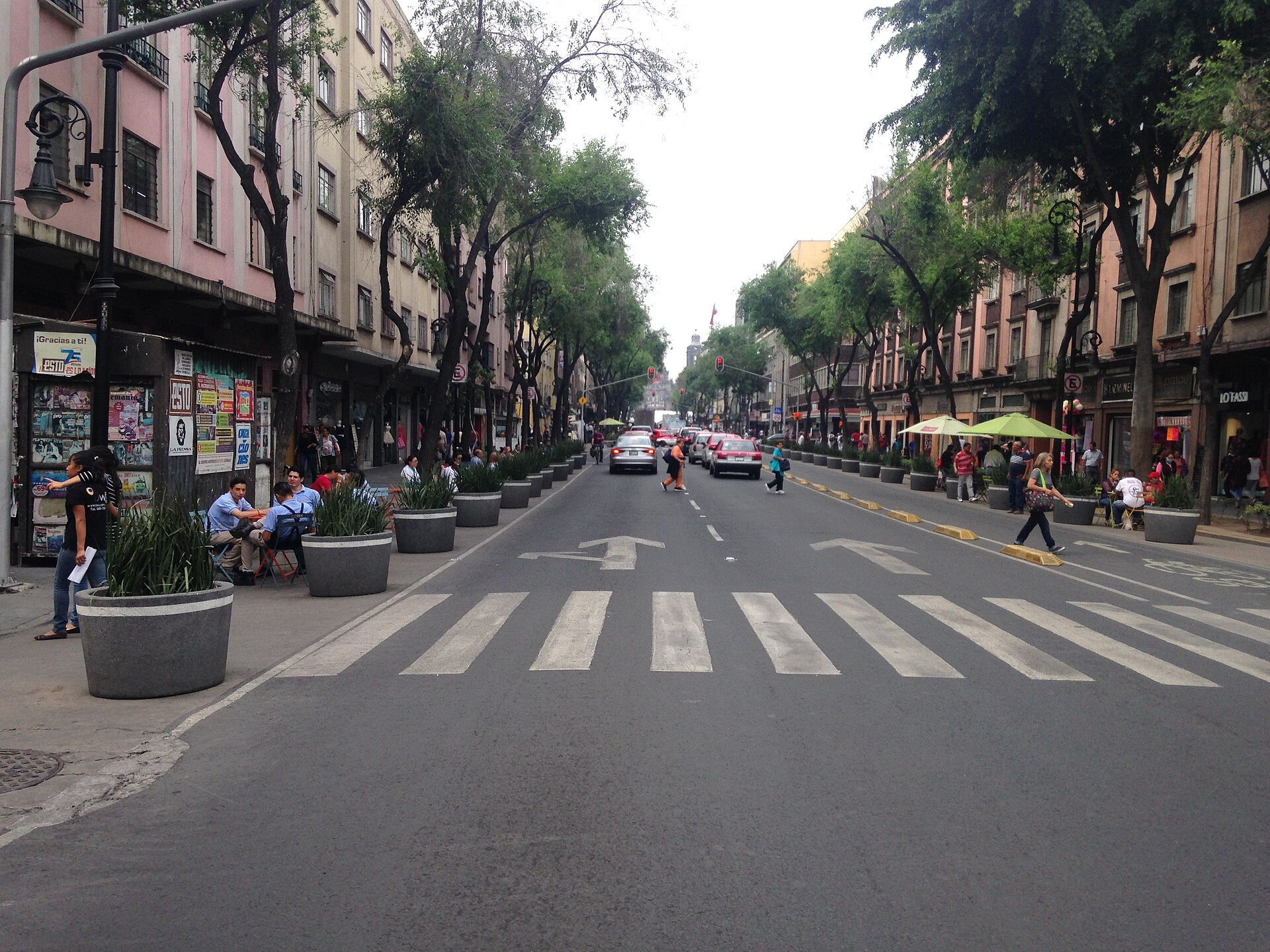 A wide tree-lined street with a zebra crossing, large planters and pedestrians in Mexico City's historic centre