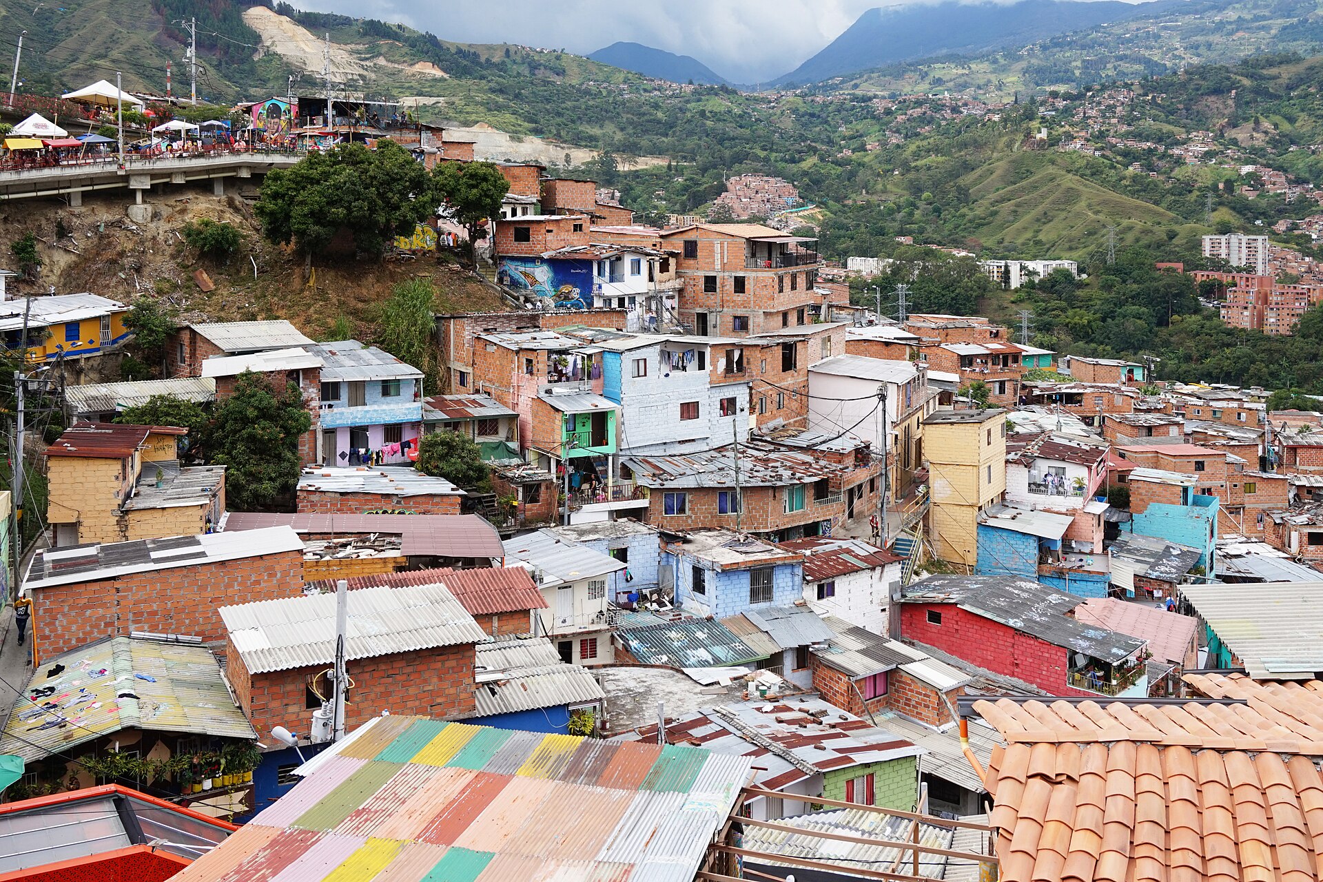 Brick and painted houses with corrugated roofs climbing a green hillside under a cloudy sky in Medellín