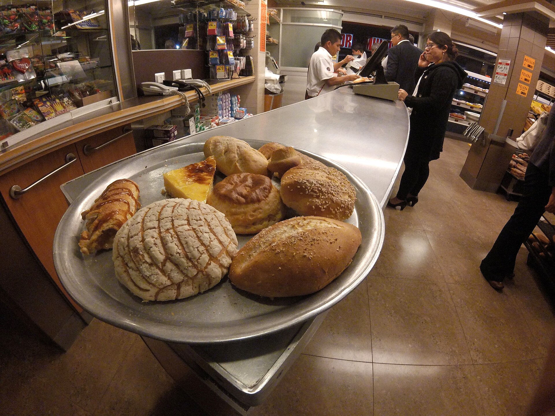 A metal tray of Mexican sweet breads on a bakery counter, including a round bun with a shell-patterned sugar crust