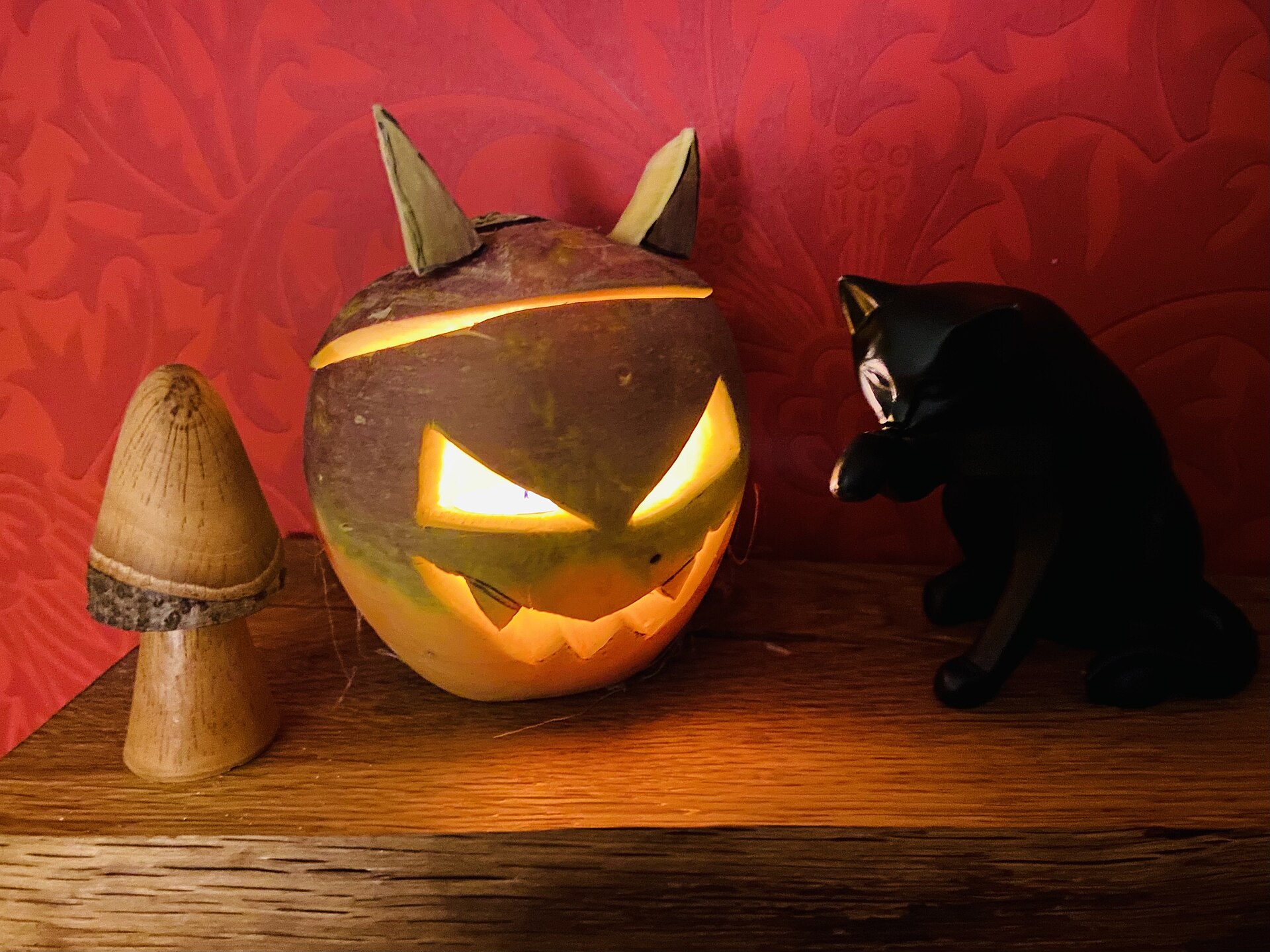 A turnip carved with a glowing jagged face and pointed ears, lit by a candle, on a wooden shelf beside a black cat figurine and a wooden toadstool