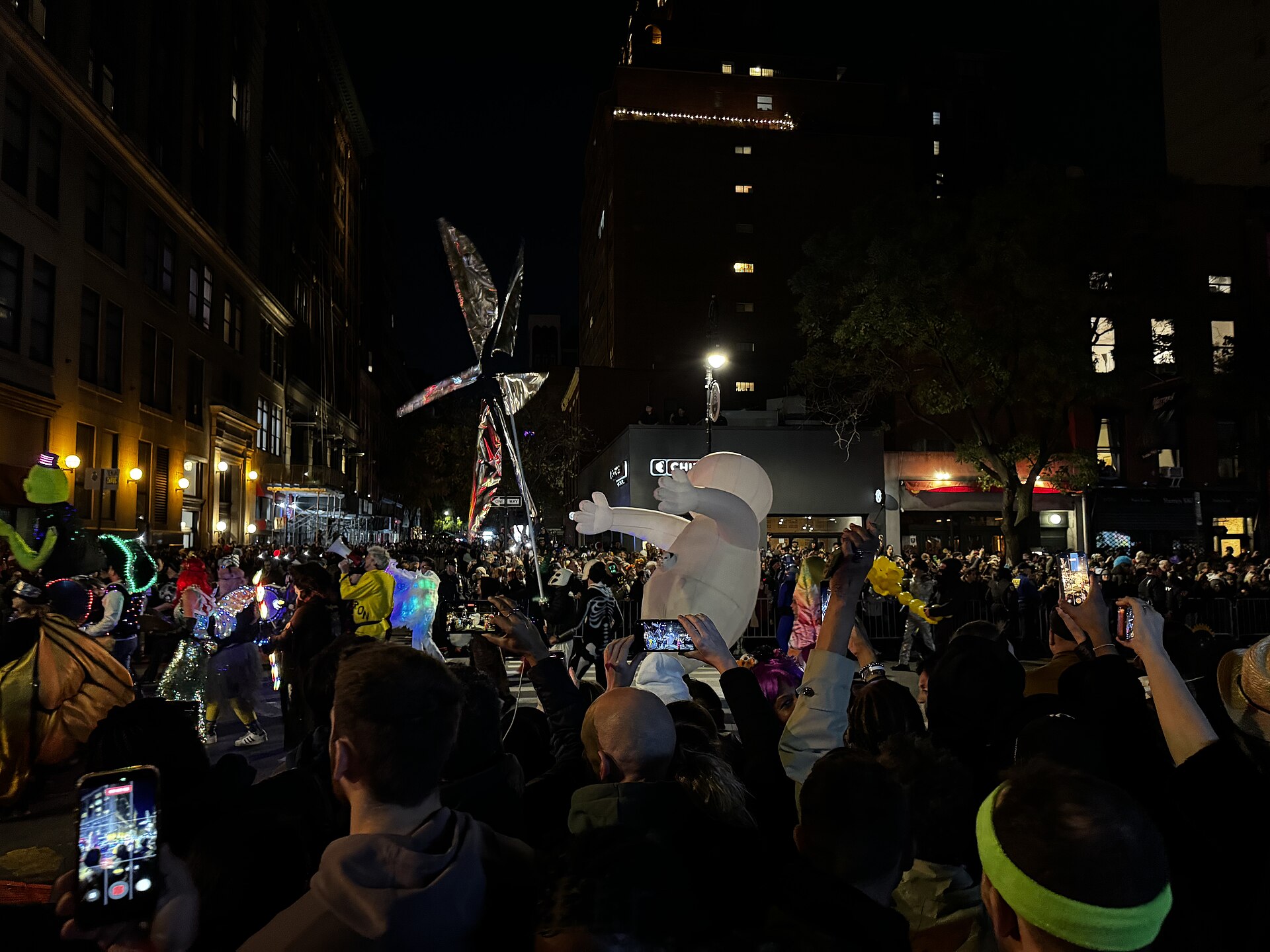 Night street scene with costumed marchers, a large white inflatable figure and a tall silver puppet, as spectators hold up phones to film