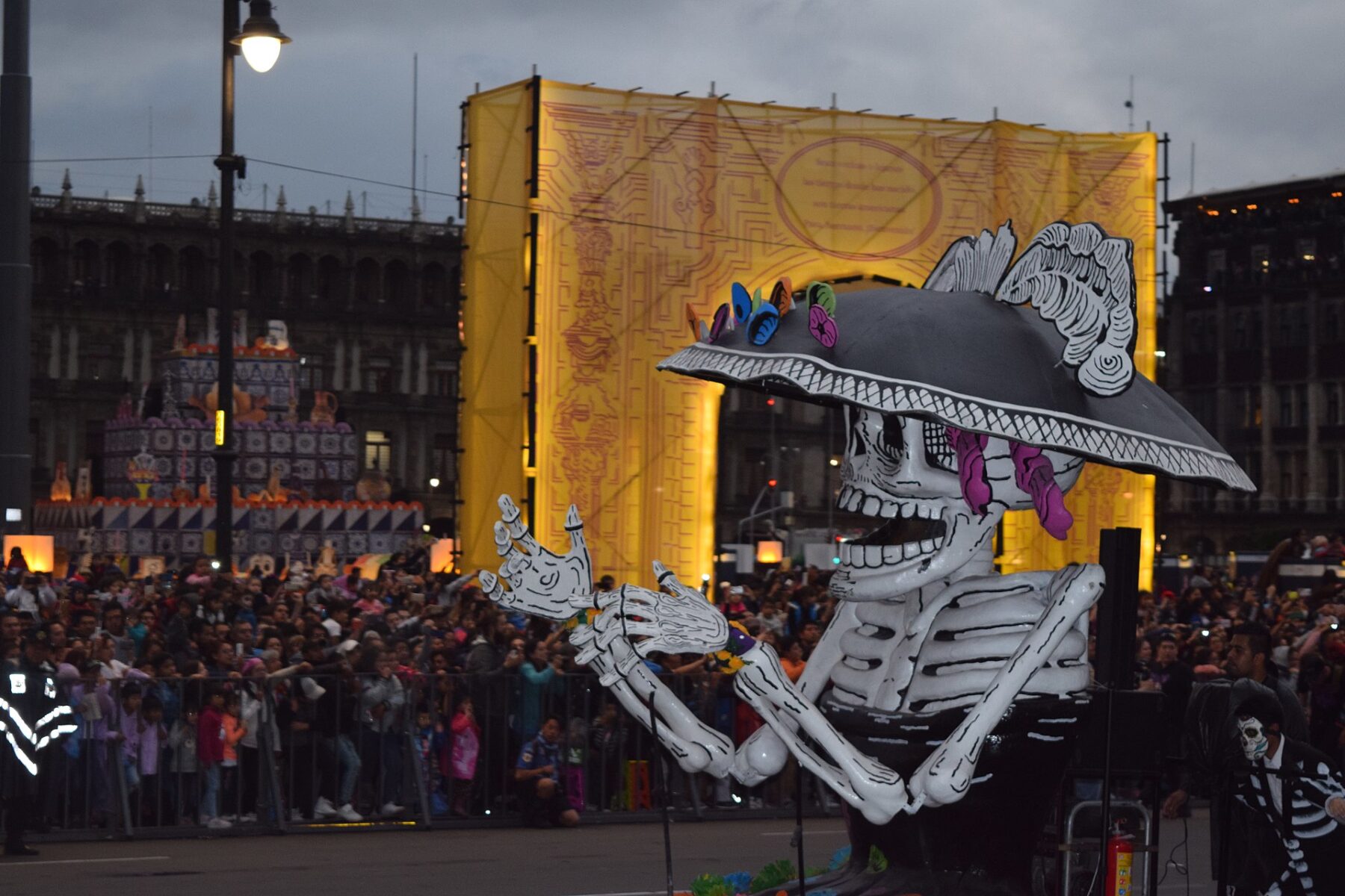 A giant skeleton float wearing a wide-brimmed hat with flowers, arms raised, passing crowds behind barriers in Mexico City's main square at dusk