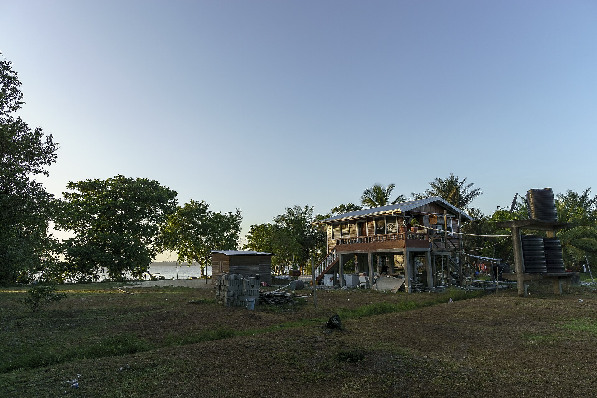 A wooden house on stilts with an outside staircase, a water tank and palm trees in an open grassy clearing near a river, in evening light
