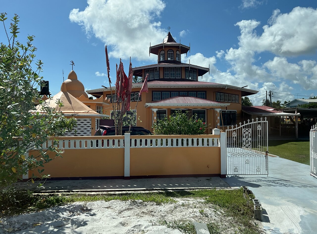 An orange Hindu temple with a tiered tower and a dome behind a low orange and white wall and gate, with tall red flags in the yard
