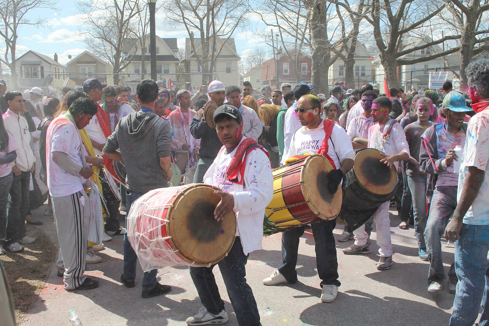 Men playing large barrel drums in a street crowd, their white clothes splashed with coloured powder, with bare trees and houses behind