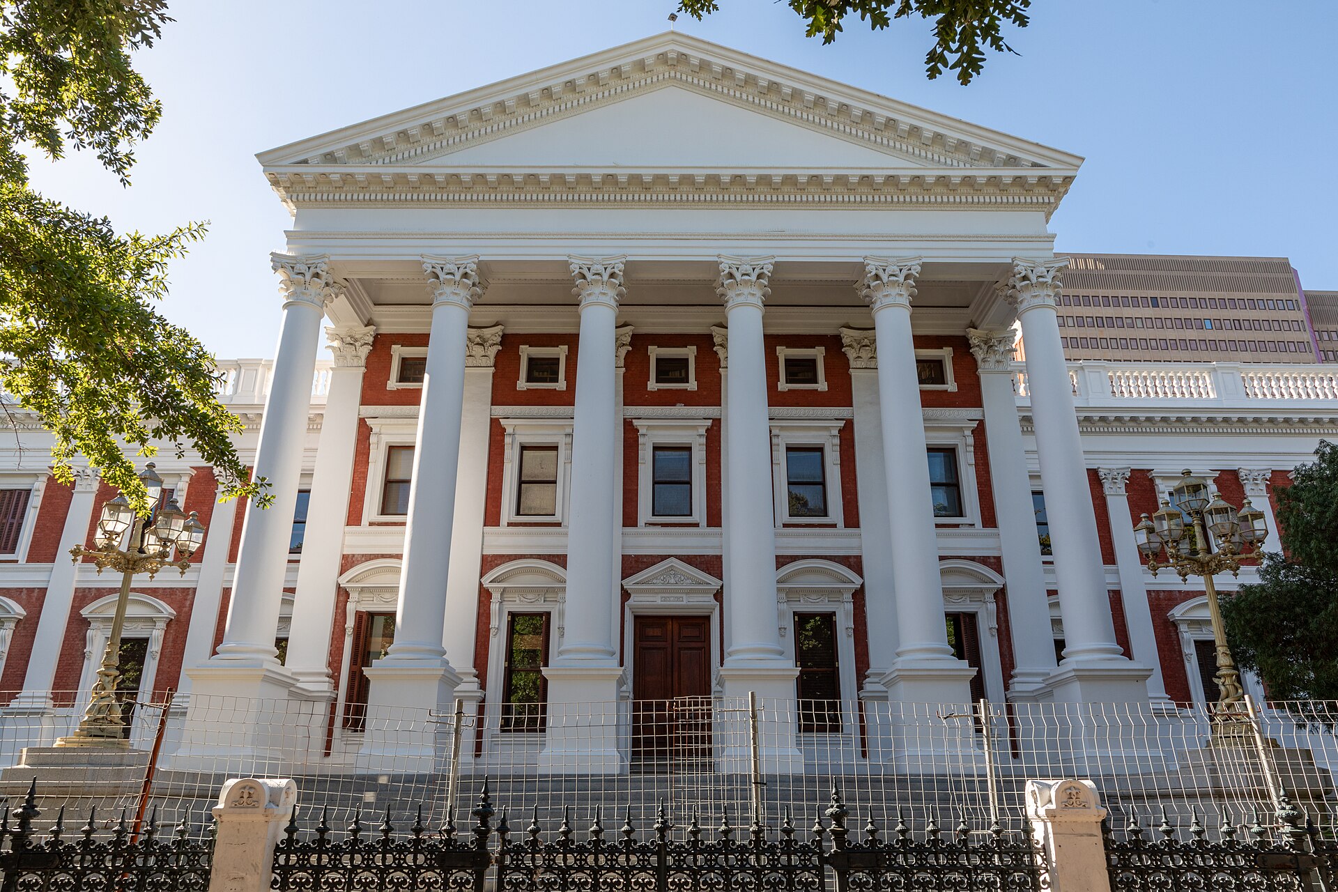 The white columned portico and red-brick facade of the Parliament of South Africa in Cape Town behind an ornate iron fence, with trees on either side