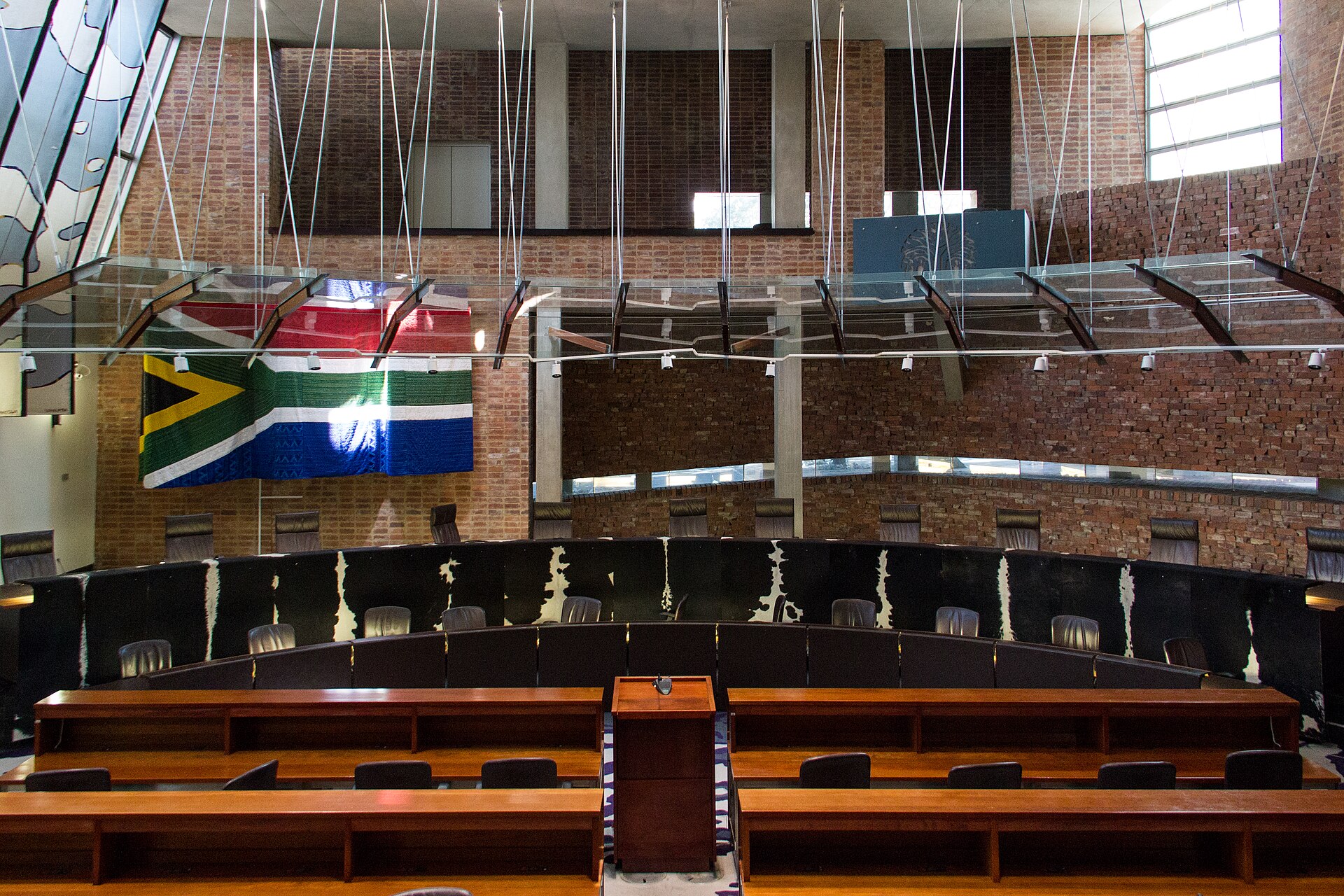 Empty courtroom of the Constitutional Court with a curved row of judges' chairs, wooden counsel tables and a large South African flag on a brick wall