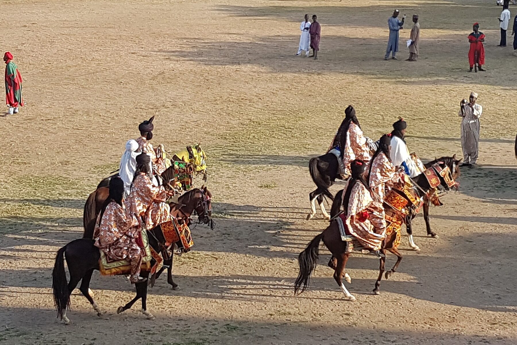 Horsemen in embroidered robes and turbans ride decorated horses across a sandy parade ground at the Durbar in Kano, with spectators in long robes standing behind them